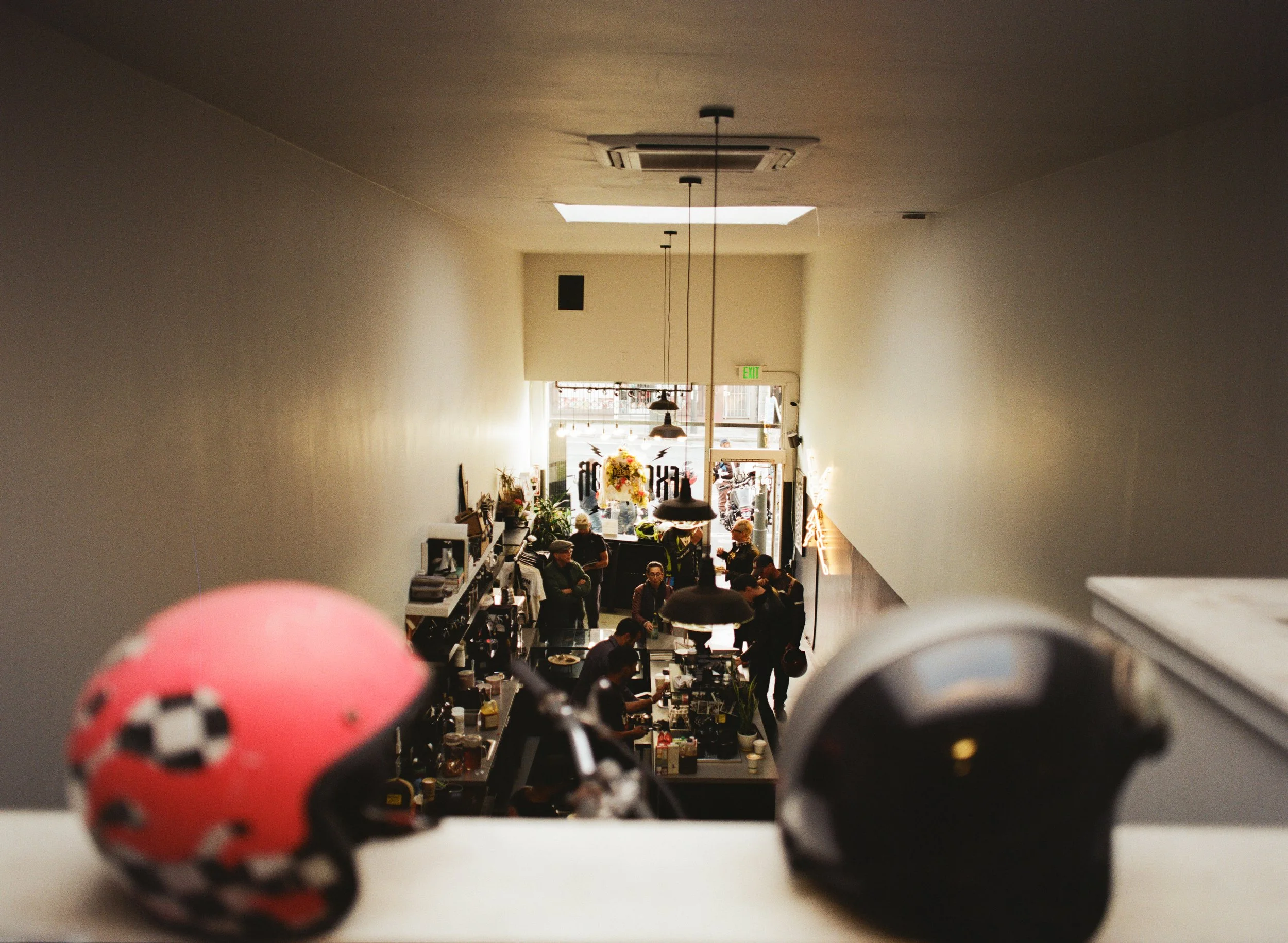 View from above showing two motorcycle helmets in the foreground and a busy cafe or restaurant with people, tables, and decor in the background.