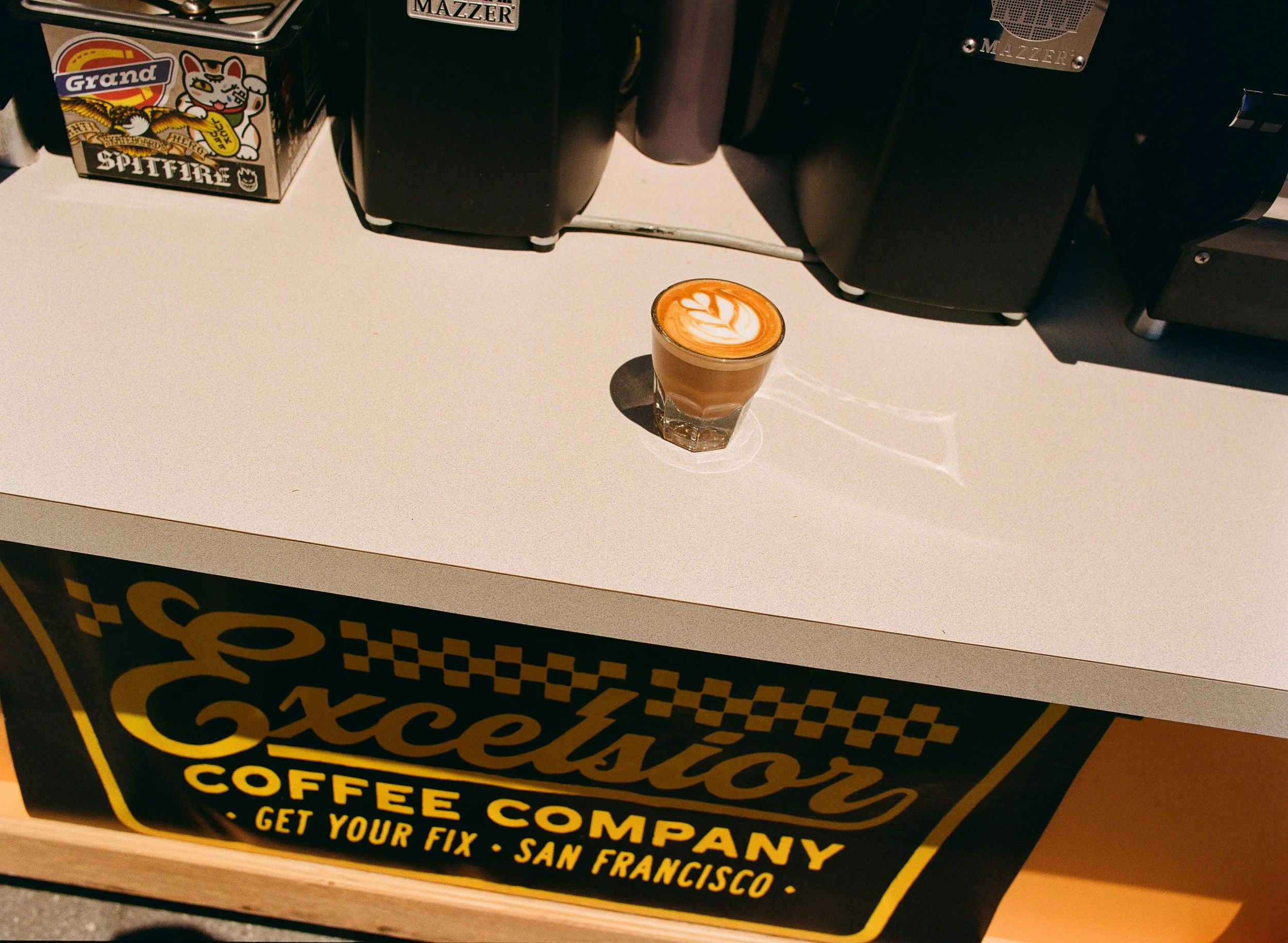 A latte with latte art on top sitting on a white countertop, with coffee machines and a box in the background, and a banner with 'Excelsior Coffee Company, Get Your Fix, San Francisco' written on it.