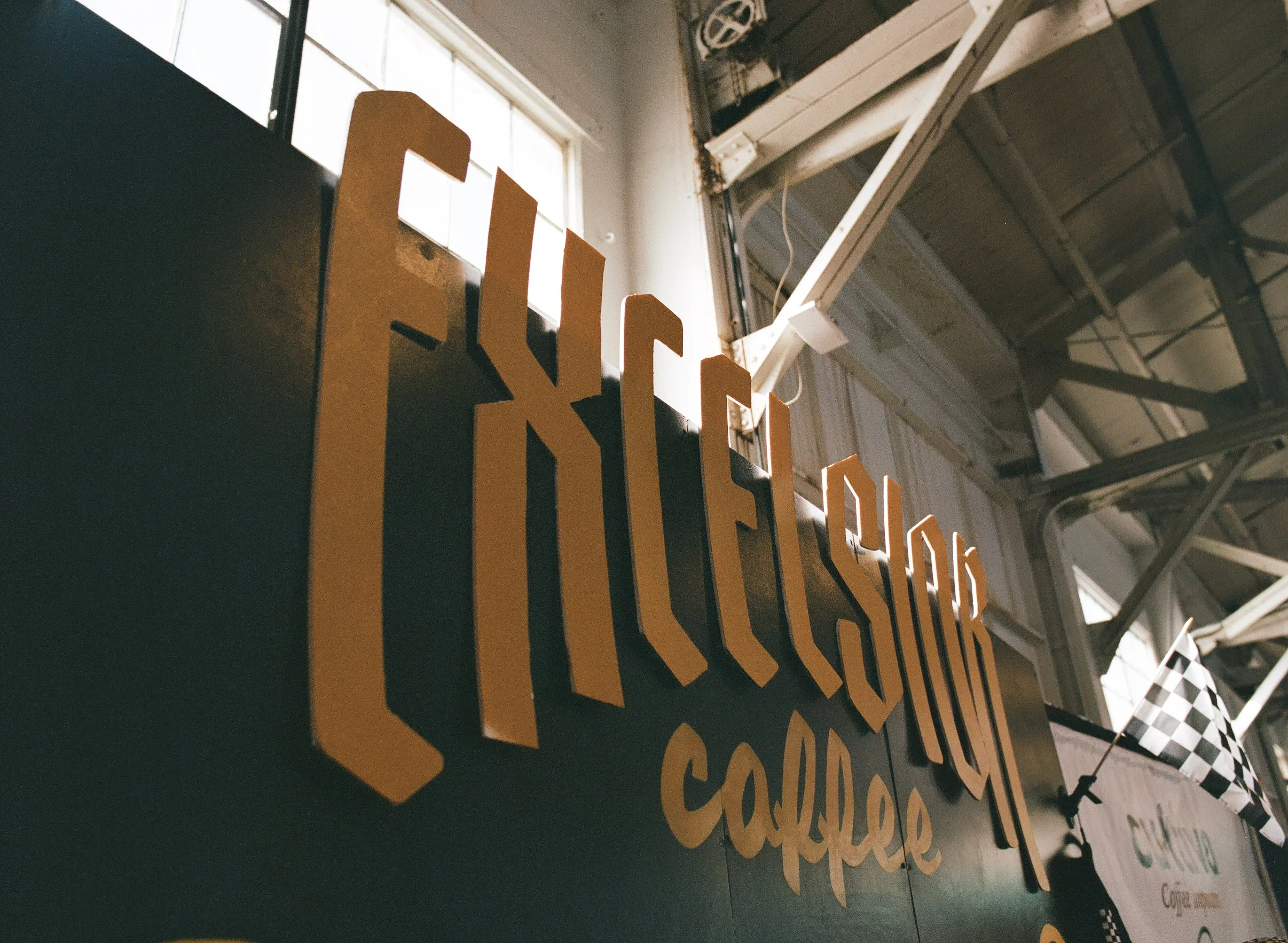 Close-up of large gold letters spelling "FUEL & FUSION coffee" on a black signboard inside a building with industrial decor, including exposed beams and windows, featuring a small checkered flag.