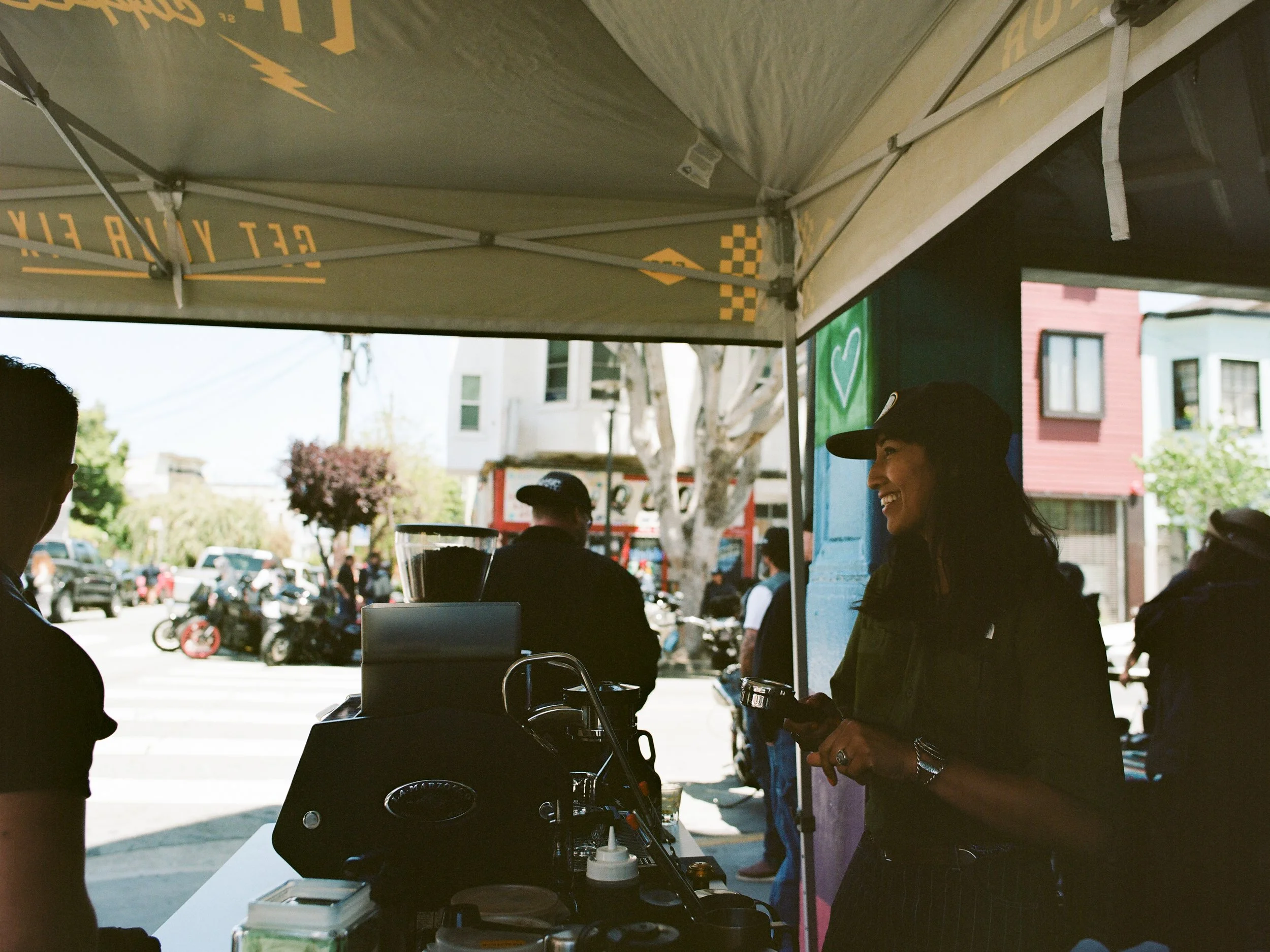 People standing and smiling under a yellow tent on a busy street, with stores and trees in the background.