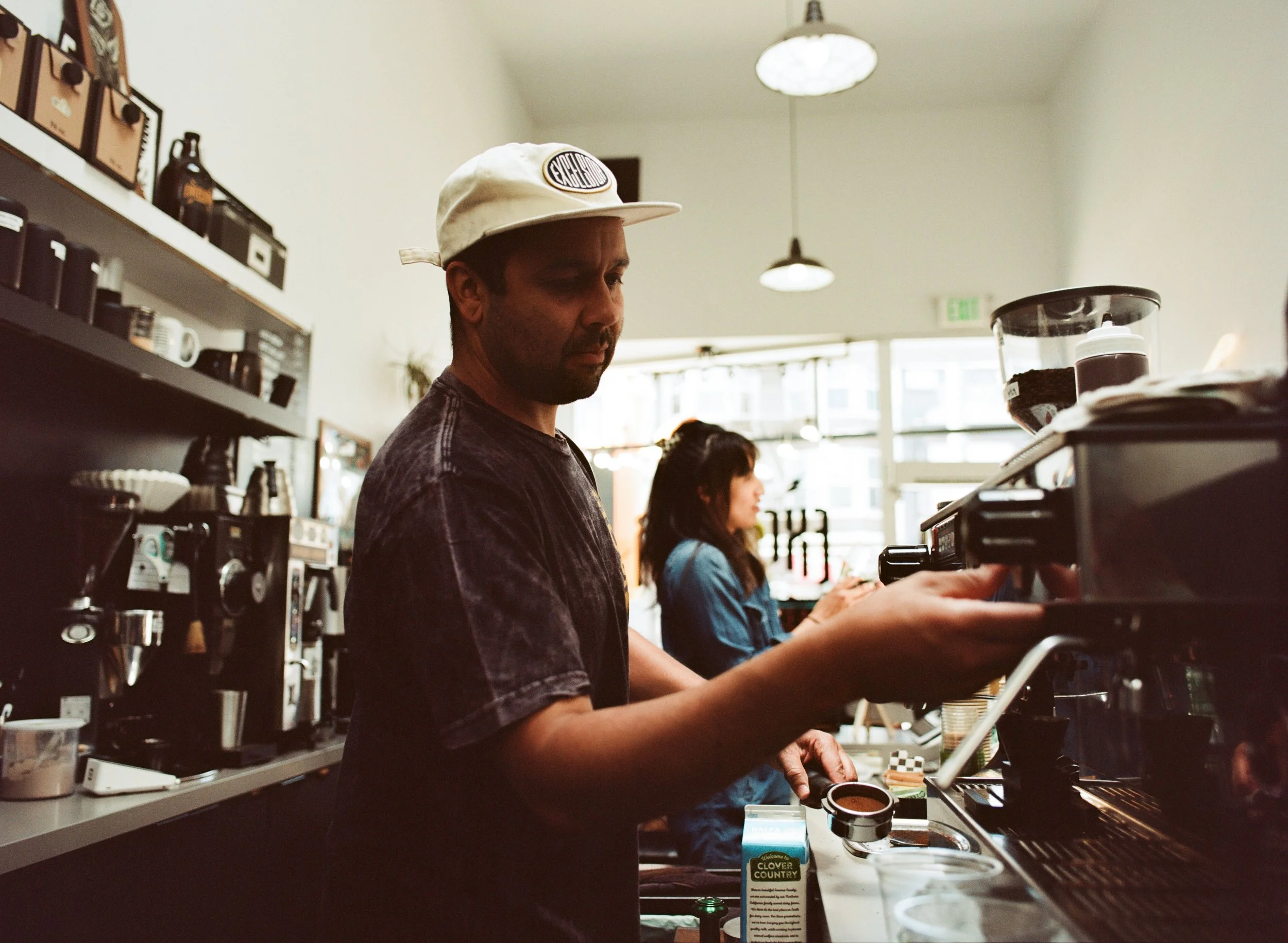 A man wearing a beige cap and black shirt making coffee behind the counter in a cafe, with a woman in a blue shirt handling a phone in the background.