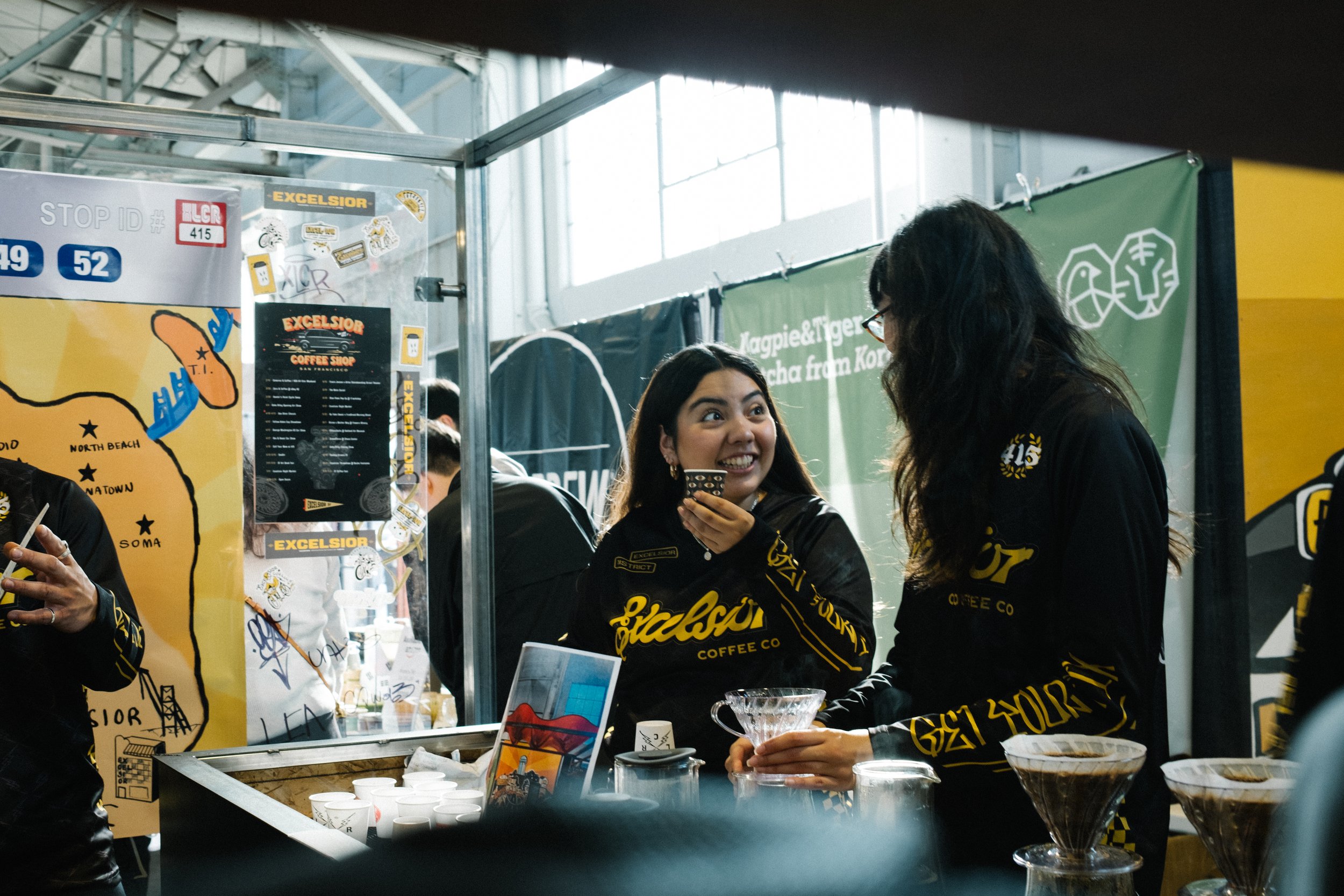 Two women wearing black jackets with yellow lettering behind a coffee stand, smiling and engaging with each other, with coffee brewing equipment and informational posters visible.