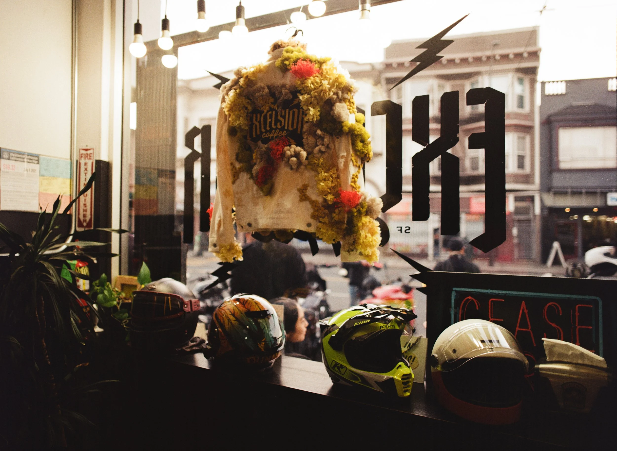 View through a cafe window shows motorcycle helmets on a counter and a floral-covered backpack hanging outside. The window has large black lettering.