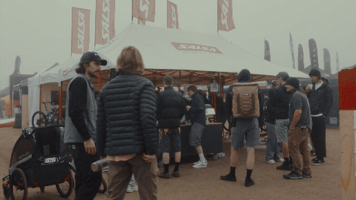 People gathered around an outdoor tent with flags, at a motorcycle event or exhibition.