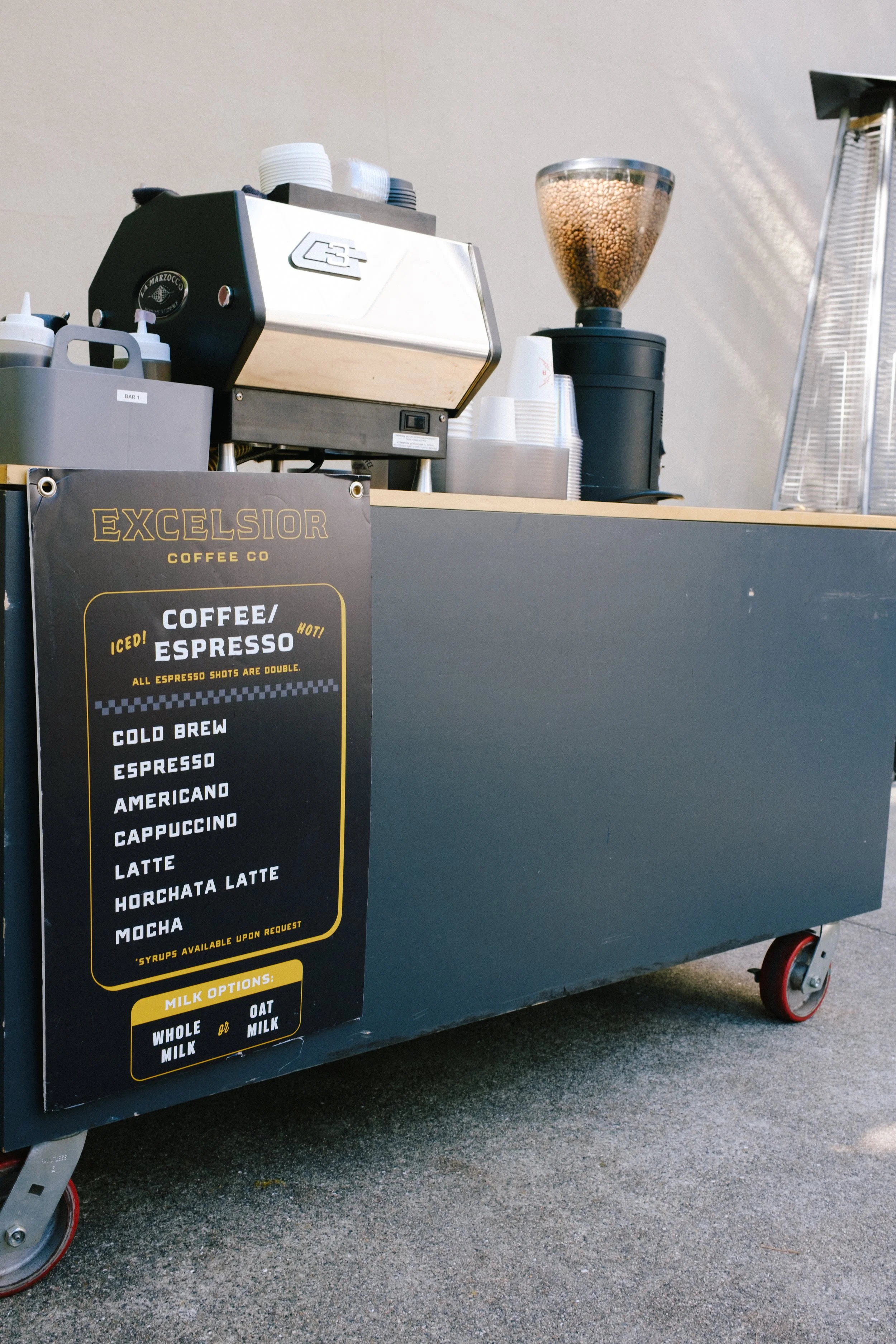 Coffee cart with a menu sign listing various coffee drinks including espresso, americano, cappuccino, latte, horchata latte, and mocha, along with milk options.