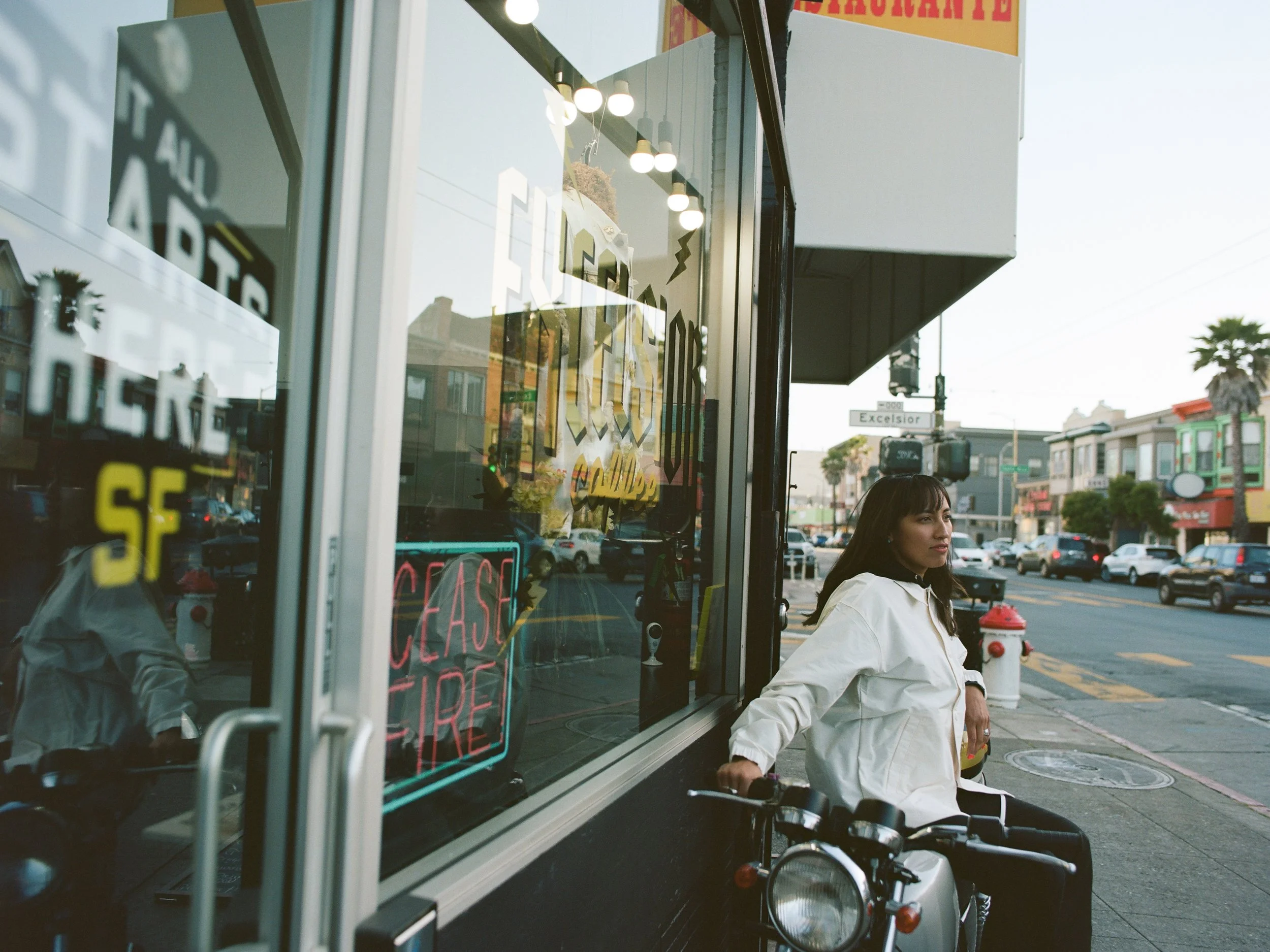 A woman with dark hair wearing a white jacket sitting on a scooter outside a storefront on a city street. The storefront has large glass windows with signs, and the street behind has cars, palm trees, and colorful buildings.