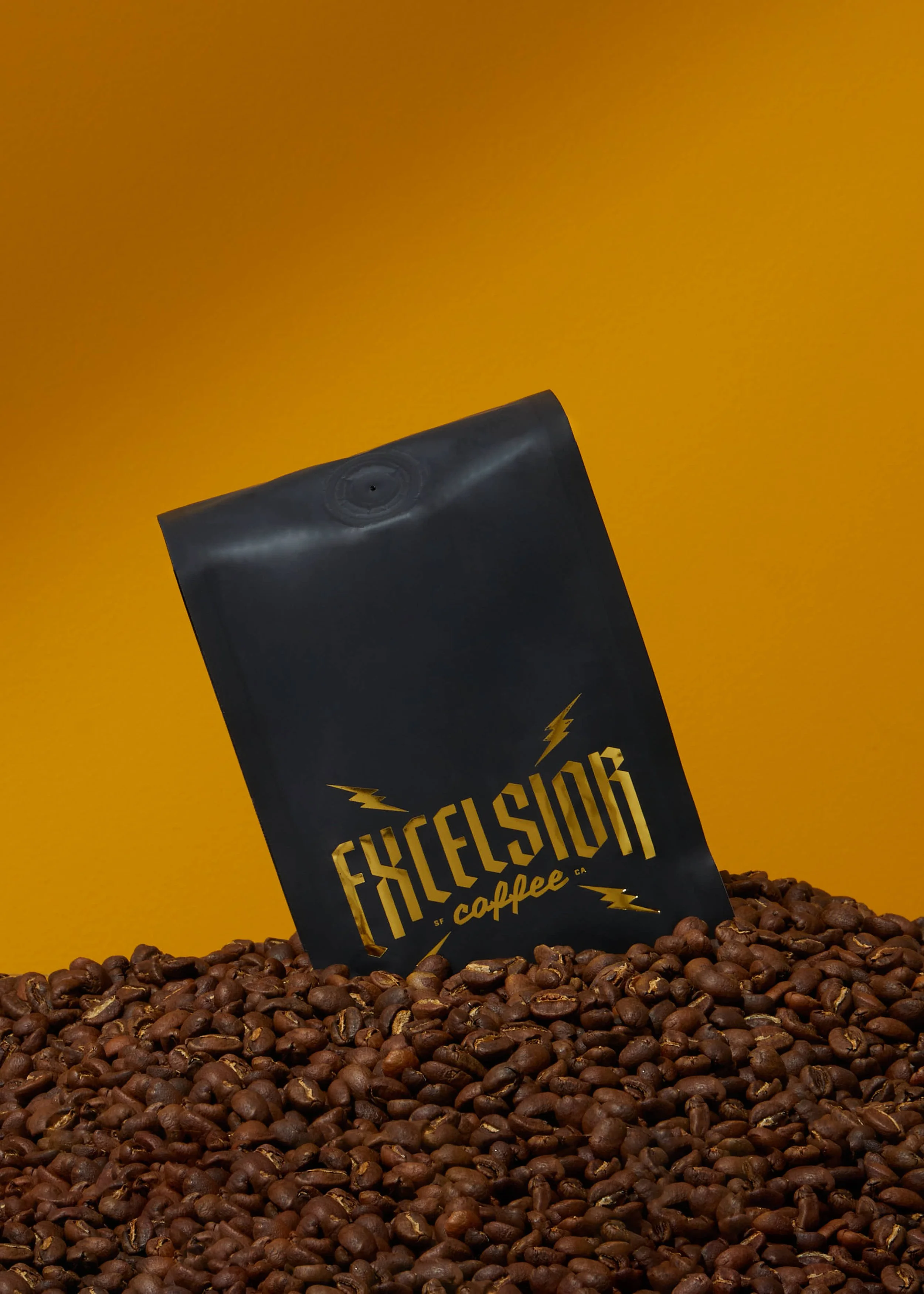 Black bag of Excelsior coffee placed among roasted coffee beans against a yellow background.