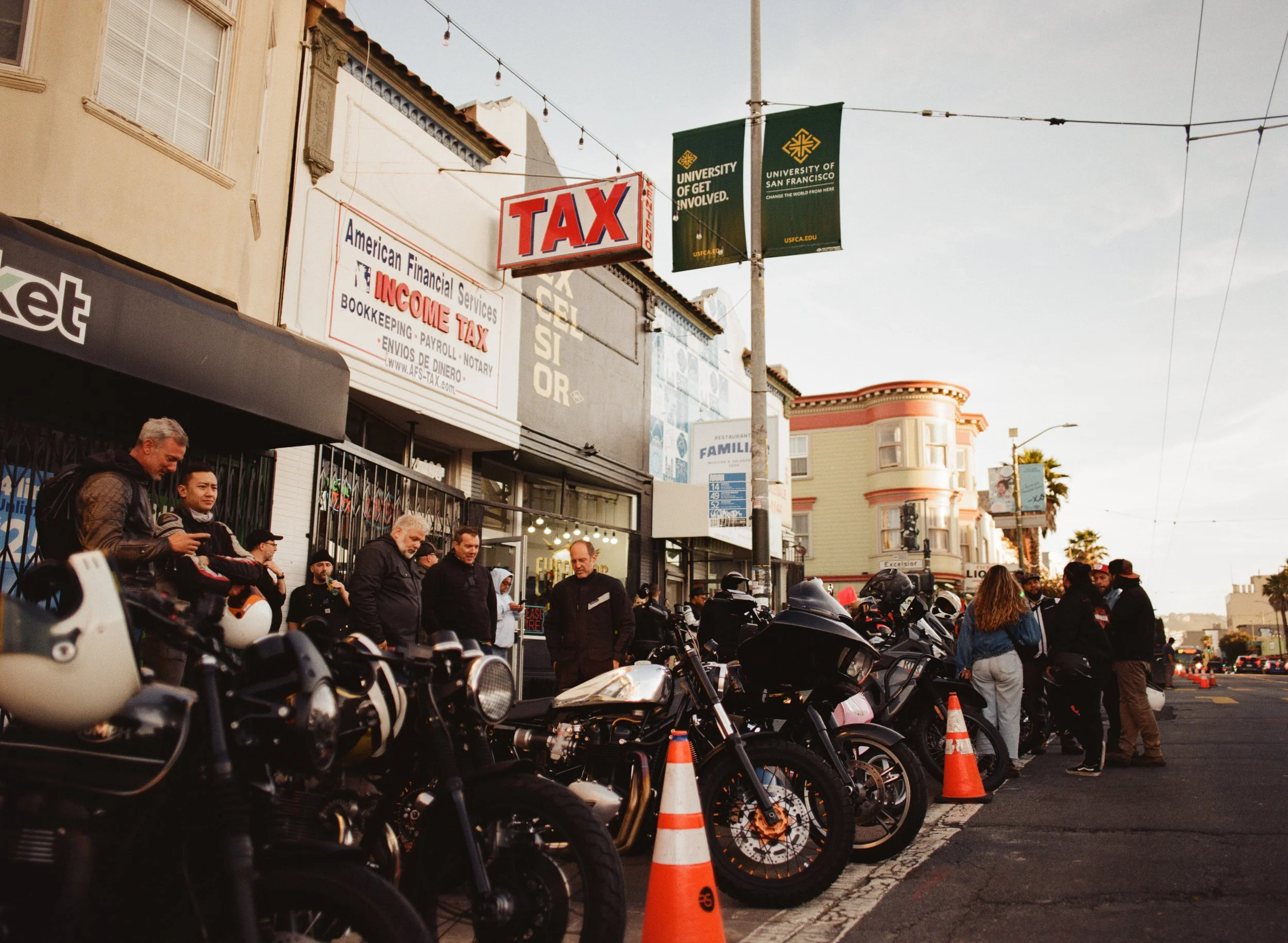 People gathered outside a storefront with motorcycles parked along the sidewalk, and traffic cones indicating a motorcycle event or gathering, in a city street at sunset.