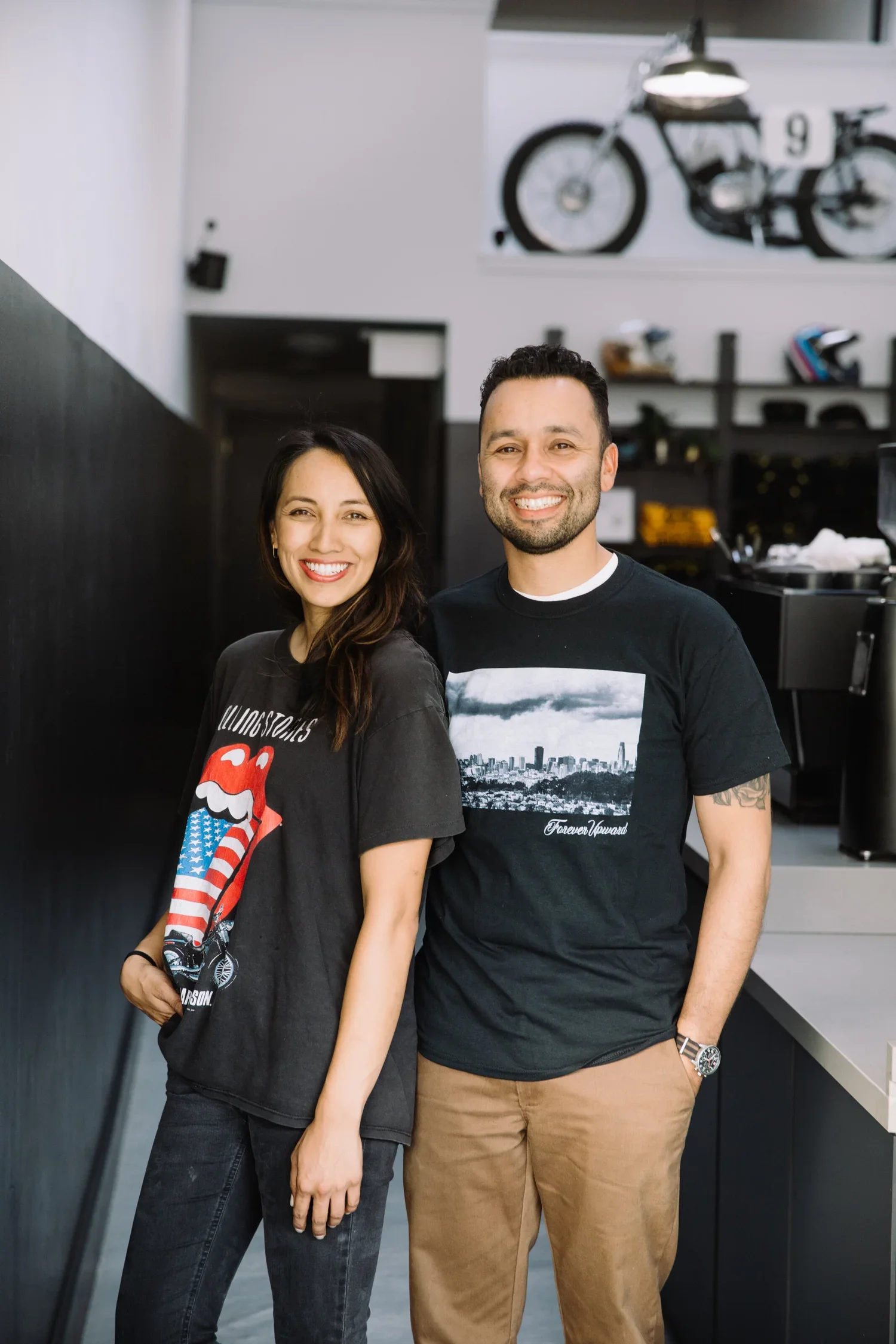 Two smiling people, a woman and a man, standing side by side in a modern indoor cafe or restaurant. In the background, there is a bicycle mounted on the wall and some shelves with items. The woman has dark hair and is wearing a black T-shirt with a Rolling Stones logo and American flag design, and the man has short dark hair, a beard, and is wearing a black T-shirt with a cityscape and gray pants.