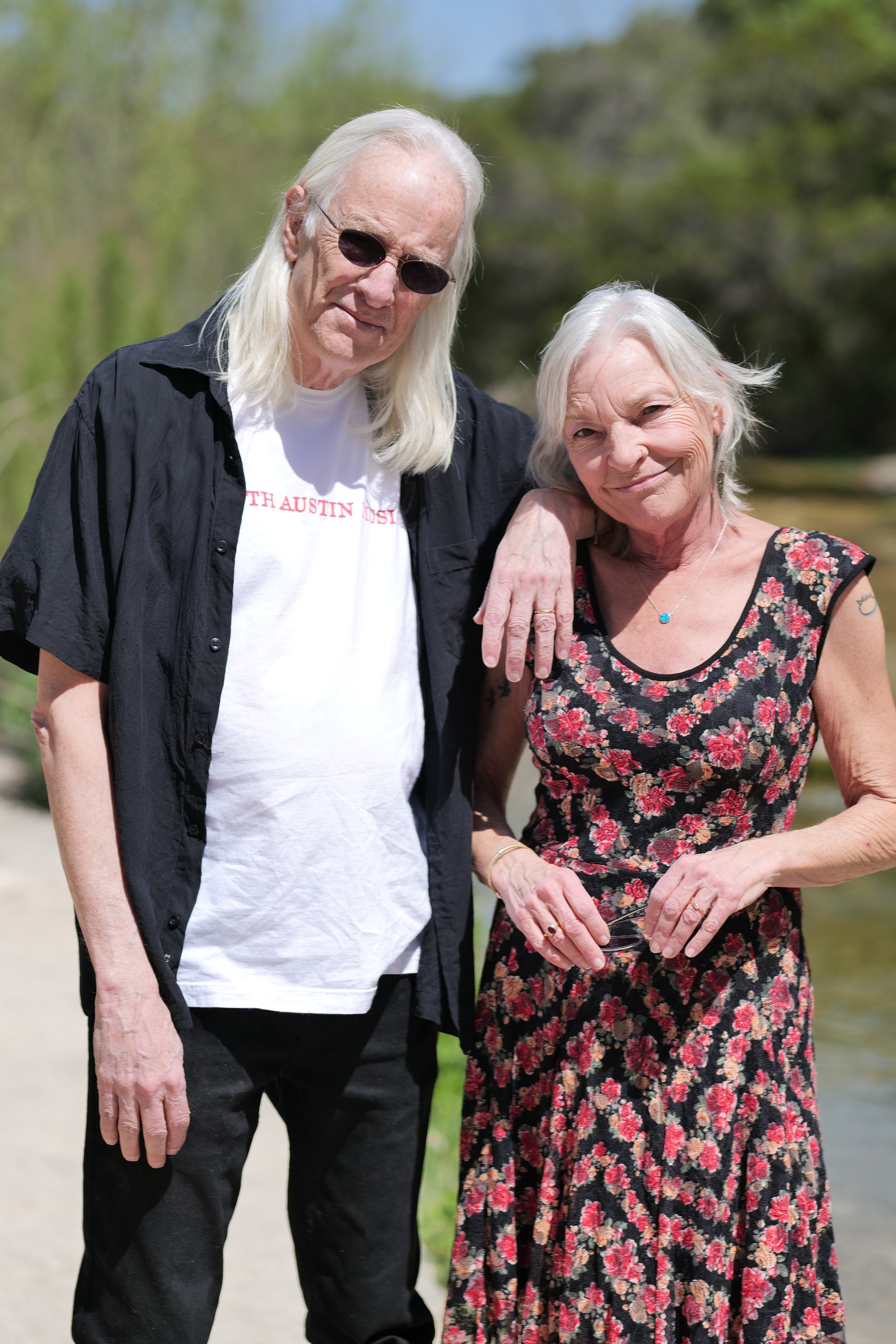 An elderly man with long white hair and sunglasses, dressed in a white T-shirt and black shirt, stands next to a smiling elderly woman with short gray hair, wearing a floral dress, outdoors near a body of water with greenery in the background.