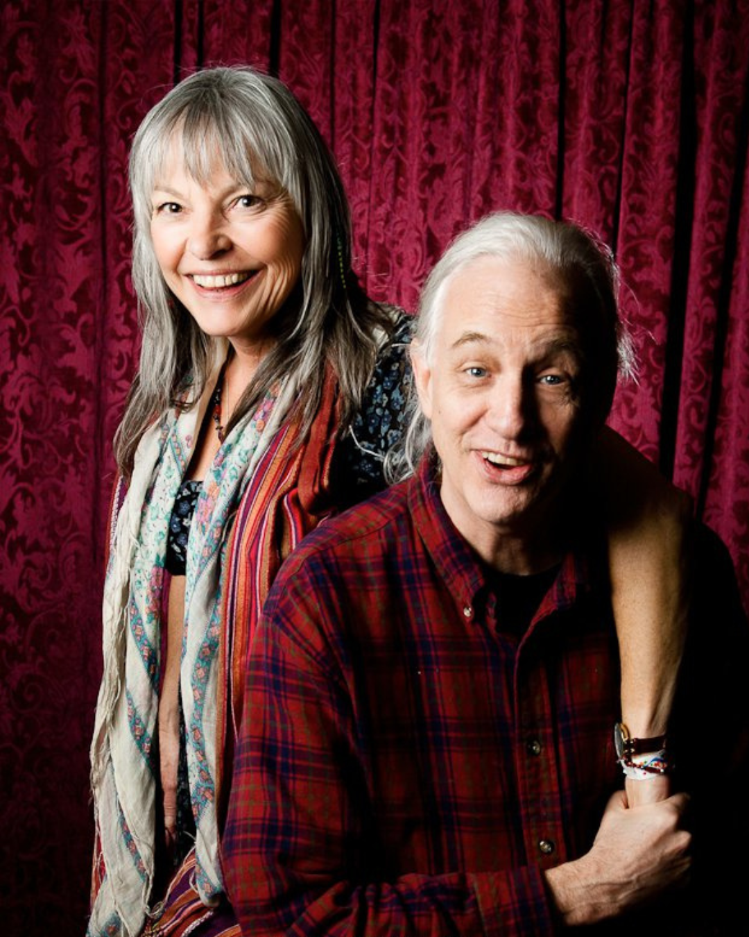 A smiling elderly woman with long gray hair and a smiling elderly man with long gray hair. The woman is standing behind and slightly to the left of the man, both are looking at the camera. They are in front of a red, floral patterned curtain.