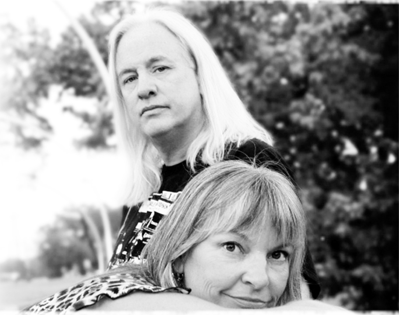Black and white photo of Jim & Sherry outdoors, one standing behind the other, with trees in the background.