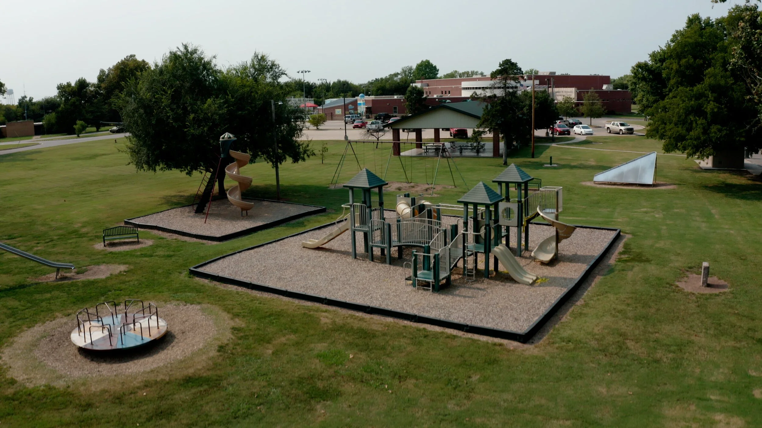 A playground with multiple play structures including slides, swings, a spiral slide, and a merry-go-round, situated on grass and surrounded by trees and a grassy field.