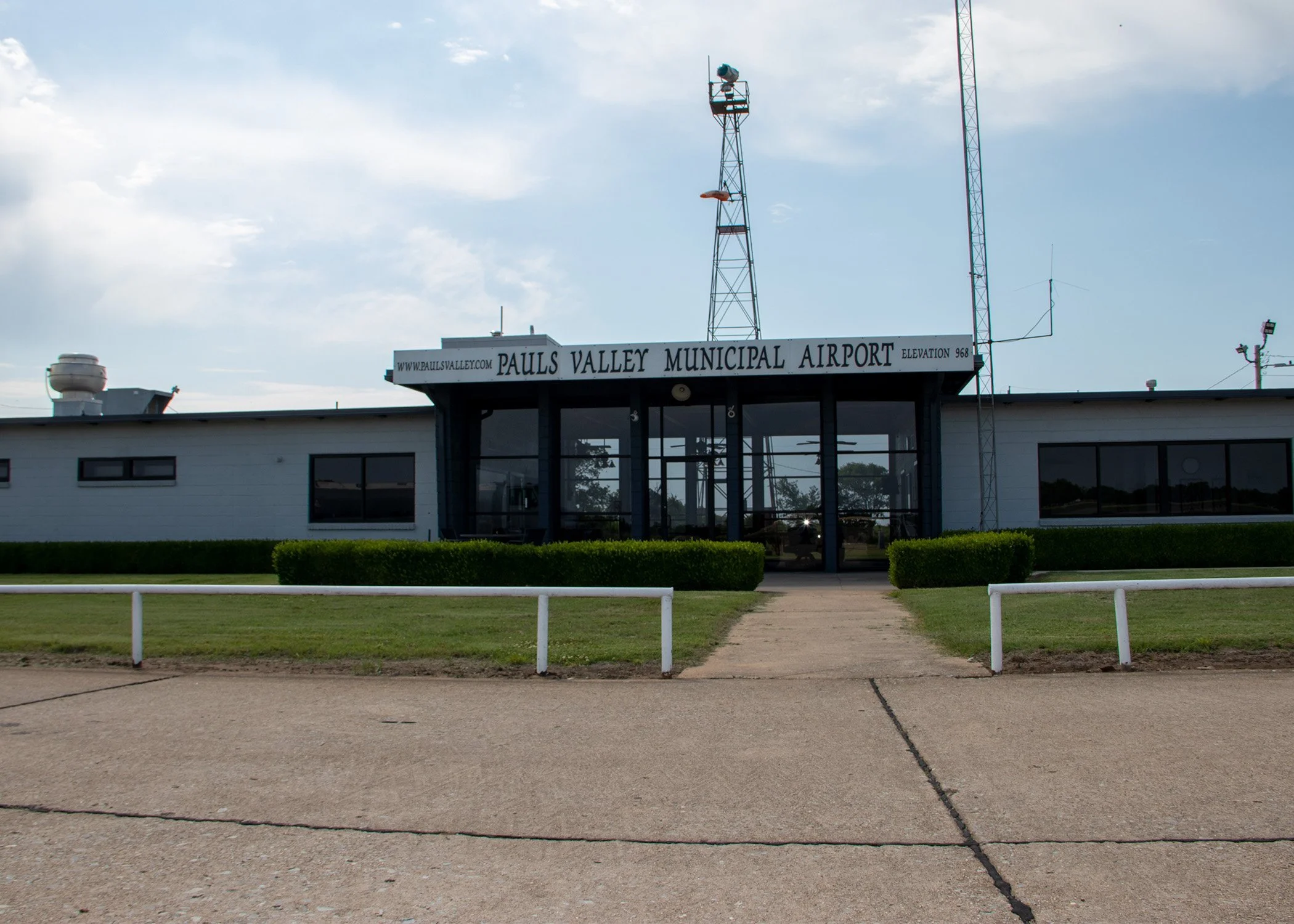 Front view of Pauls Valley Municipal Airport with building, entrance walkway, green bushes, and an antenna tower overhead.