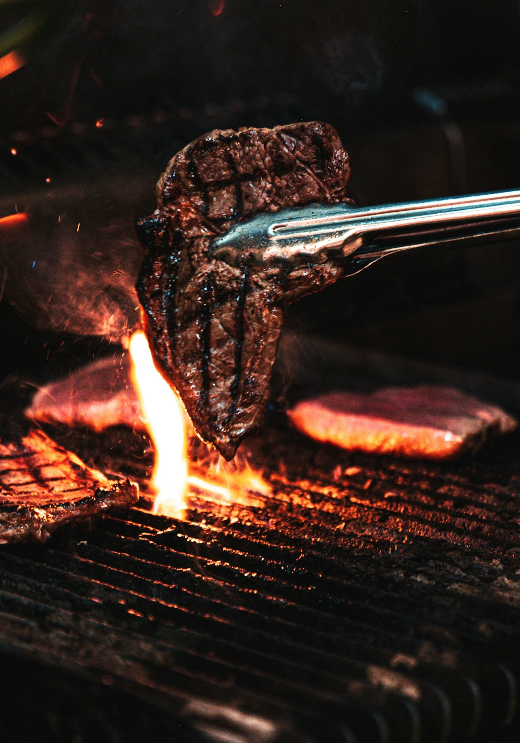 A piece of steak being cooked on a grill over open flame using tongs.