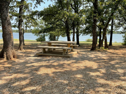 Picnic table and benches in a shaded lakeside park with trees and water in the background.