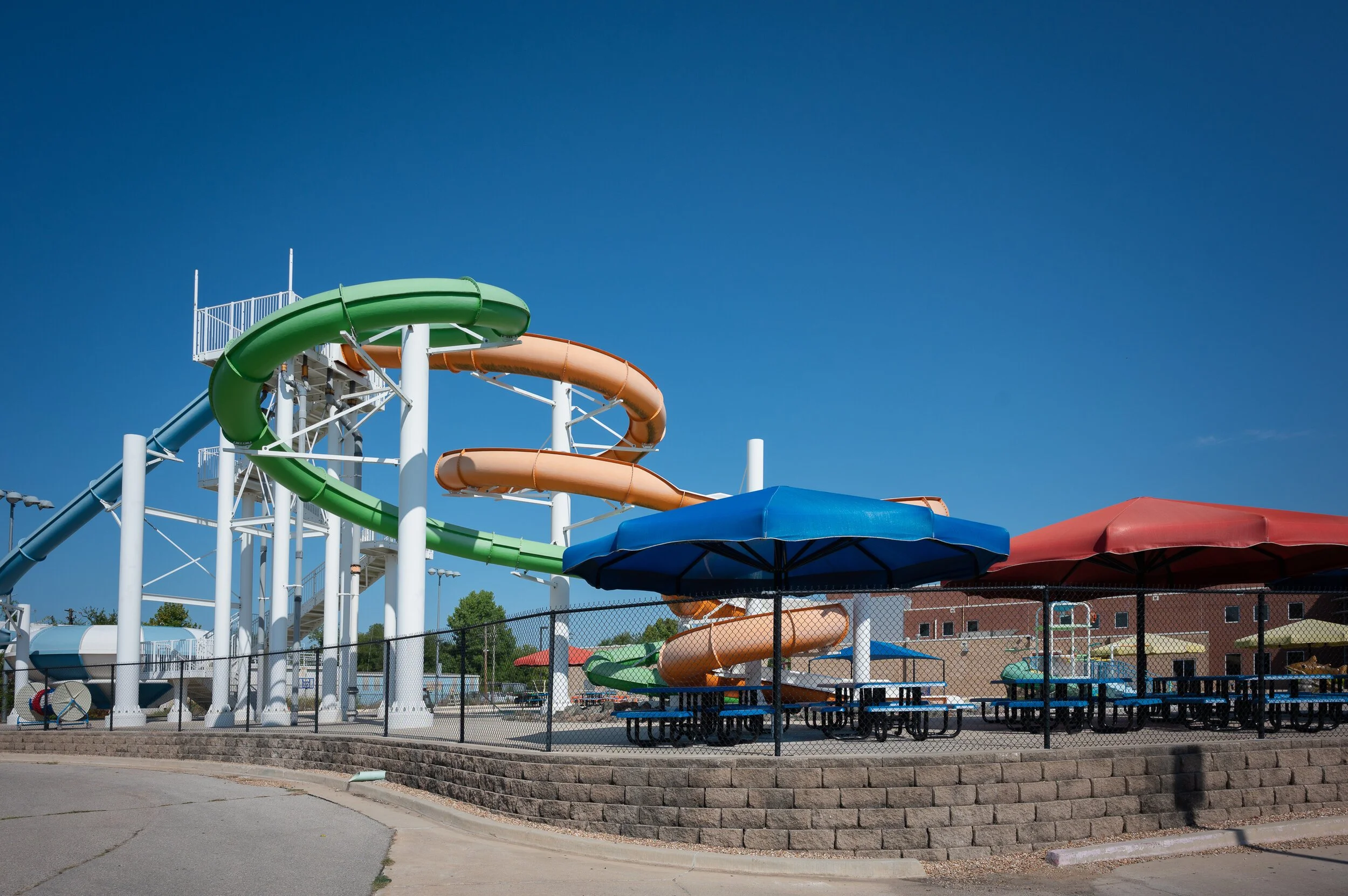 A water park with multiple water slides in green, orange, and blue, under blue and red umbrellas, surrounded by a black fence and a brick retaining wall, with buildings in the background and a clear blue sky overhead.