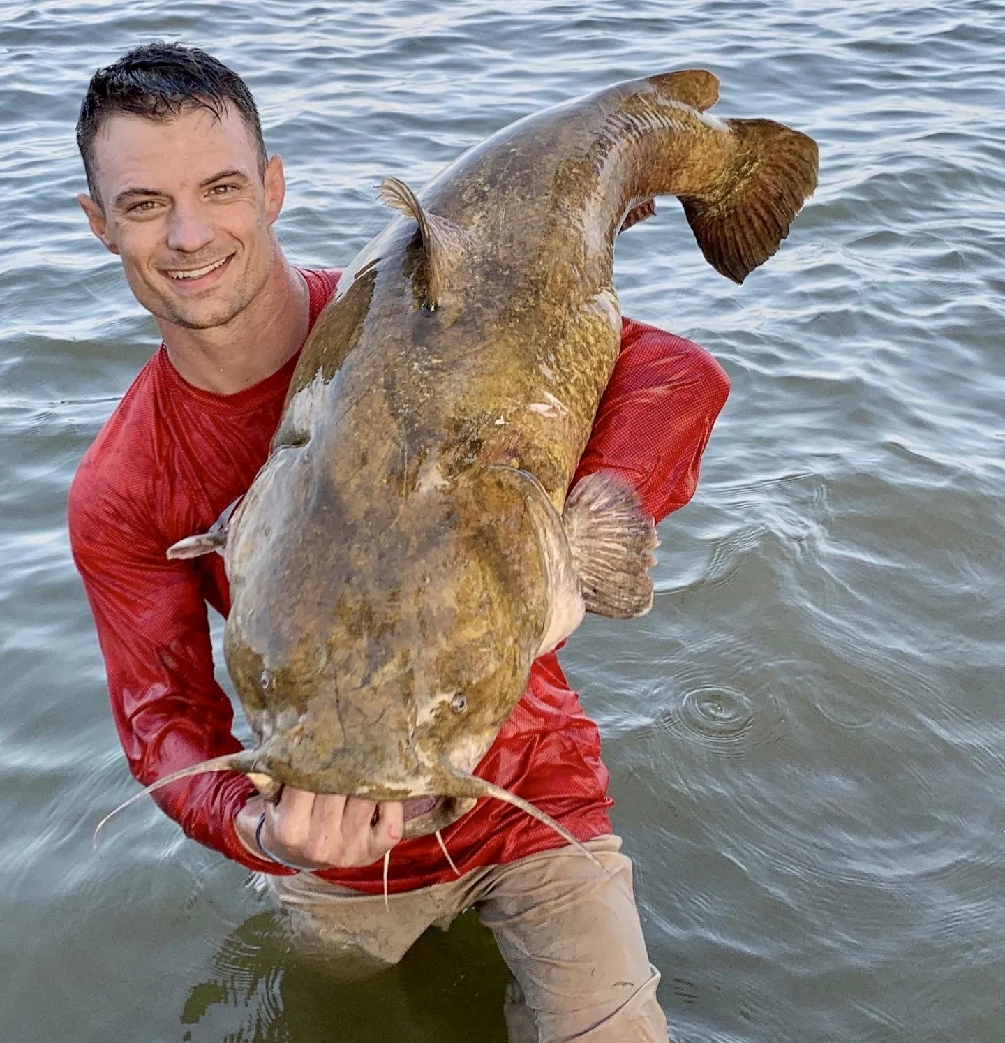 A man in a red shirt holding a large catfish over water, smiling at the camera.