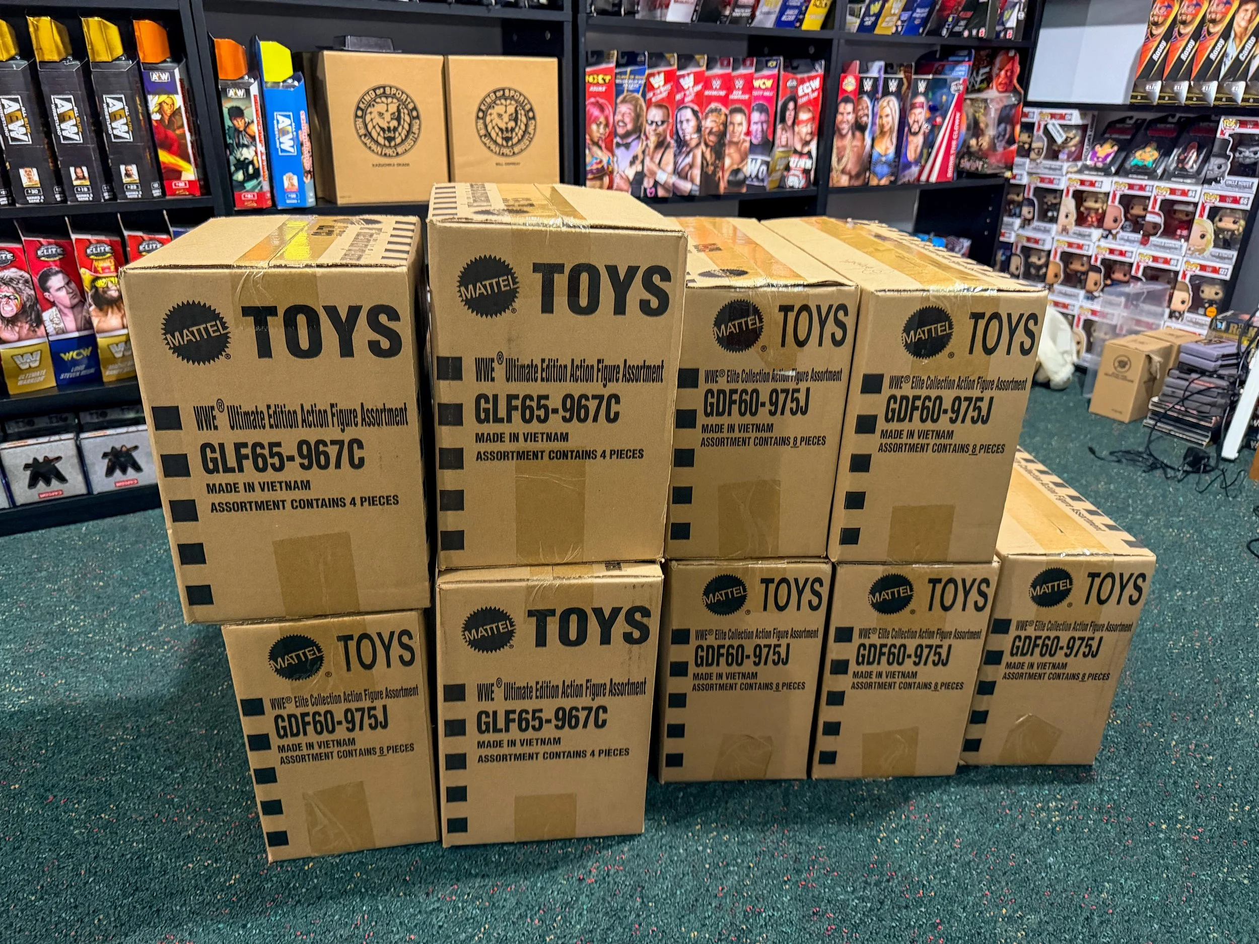 Stack of WWE Ultimate Edition action figure boxes on a store shelf, with shelves of wrestling-themed toys and collectibles in the background.