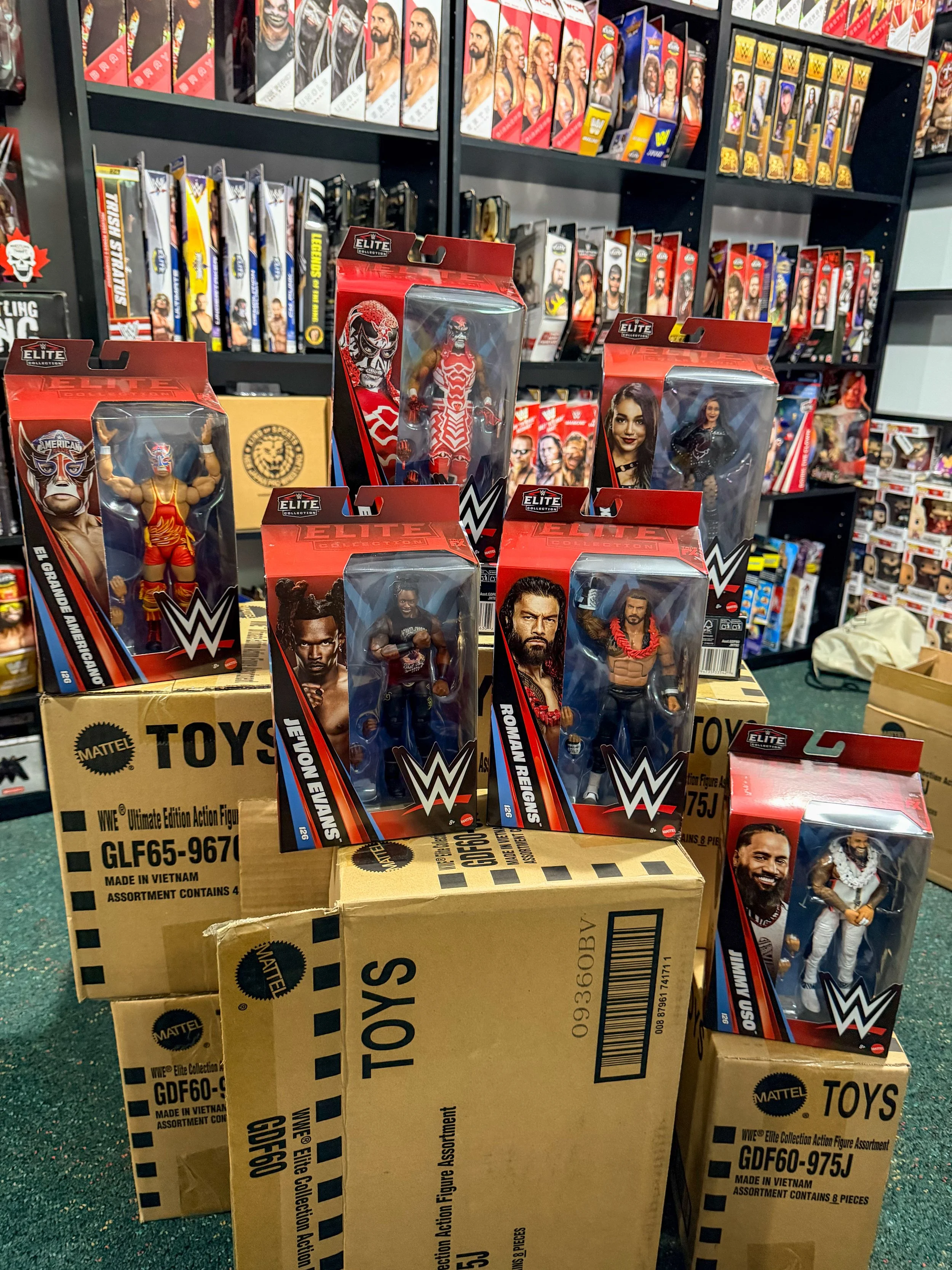 A display of WWE action figures on a table with a background of shelves filled with WWE action figure boxes. The figures include various wrestlers, some in costumes and some with realistic looks.