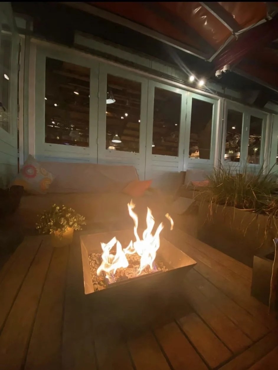 Gemütliche Veranda mit Feuerstelle, bequemen Sofas, Pflanzen und Blick durch große Fenster in eine rustikale Inneneinrichtung bei gedämpftem Licht.