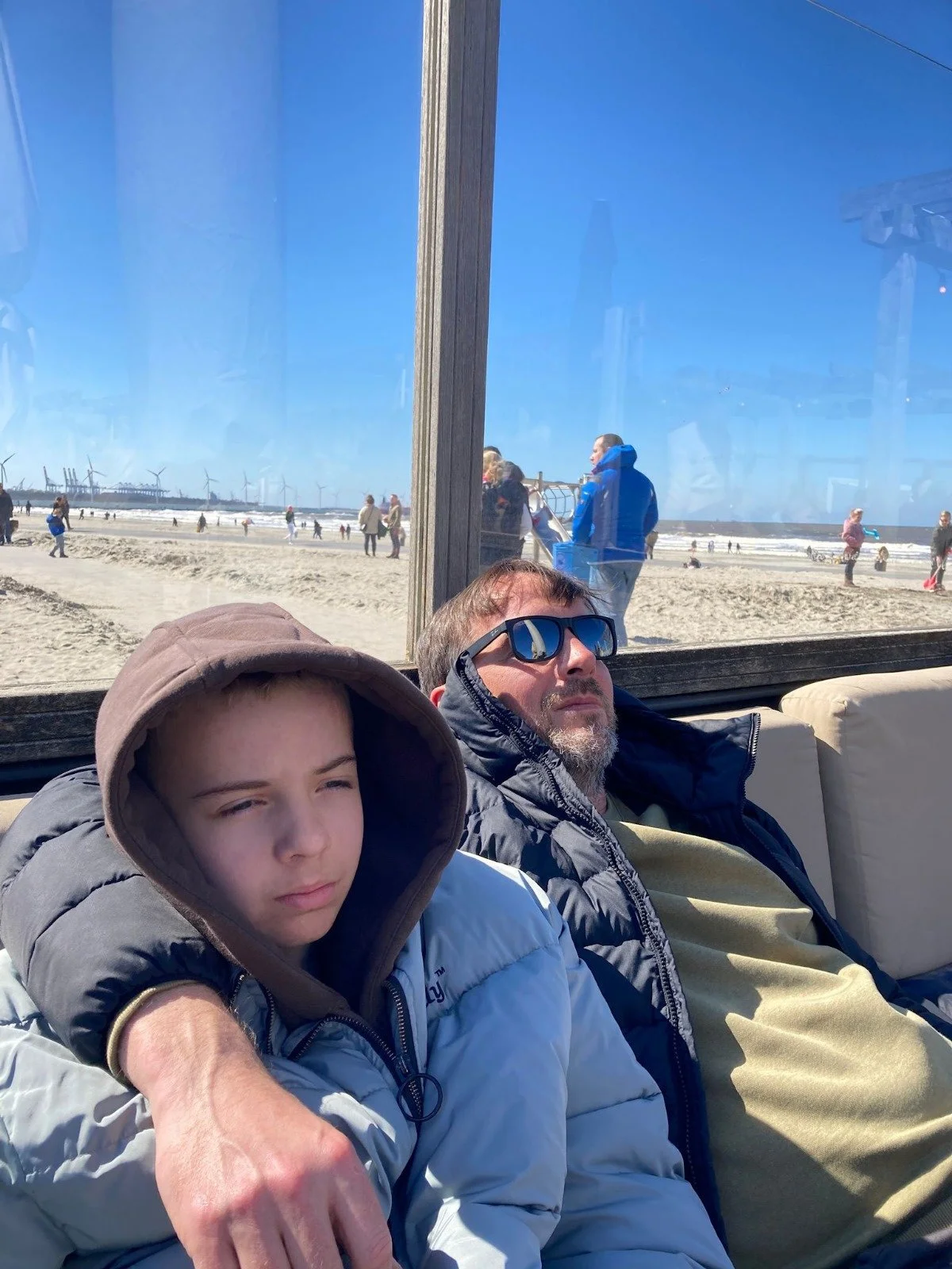 Zwei Jungen sitzen entspannt in einem Strandcafé, hinter einer großen Fensterscheibe mit Blick auf den Strand und den blauen Himmel. Es sind einige Menschen am Strand und im Hintergrund drehen Windkraftanlagen.
