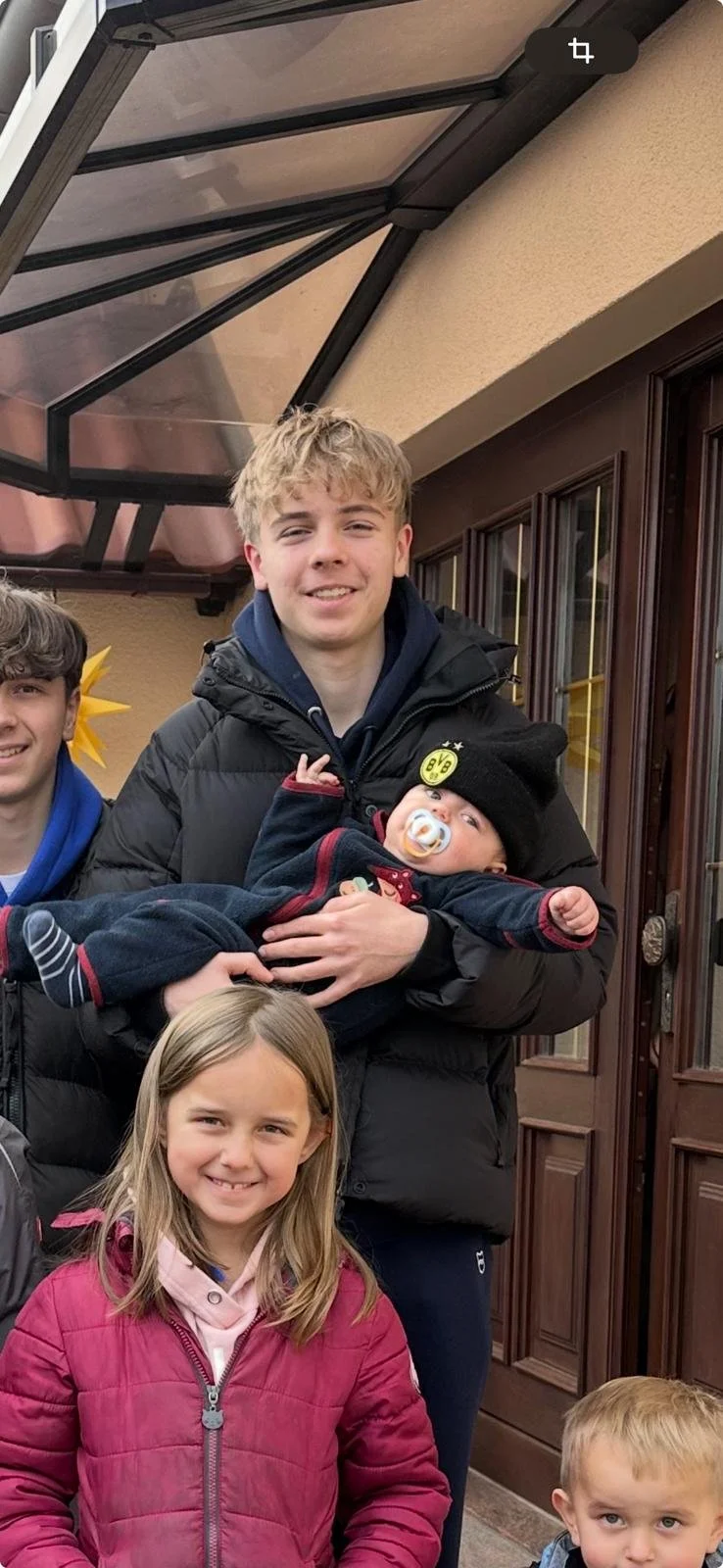 Gruppe von fünf Kindern vor einem Haus, eines hält ein Baby, alle lächeln.