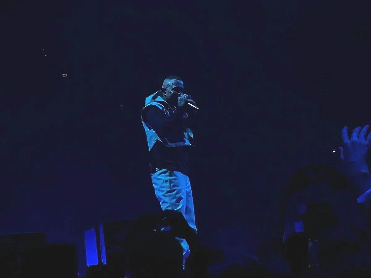 Finn war beim Luciano Konzert in Köln in der Lanxess Arena 2024. Ein junger Mann trägt eine schwarze Jacke und weiße Hosen, singt oder spricht in ein Mikrofon, während er auf einer dunklen Bühne bei einer Veranstaltung oder einem Konzert steht.