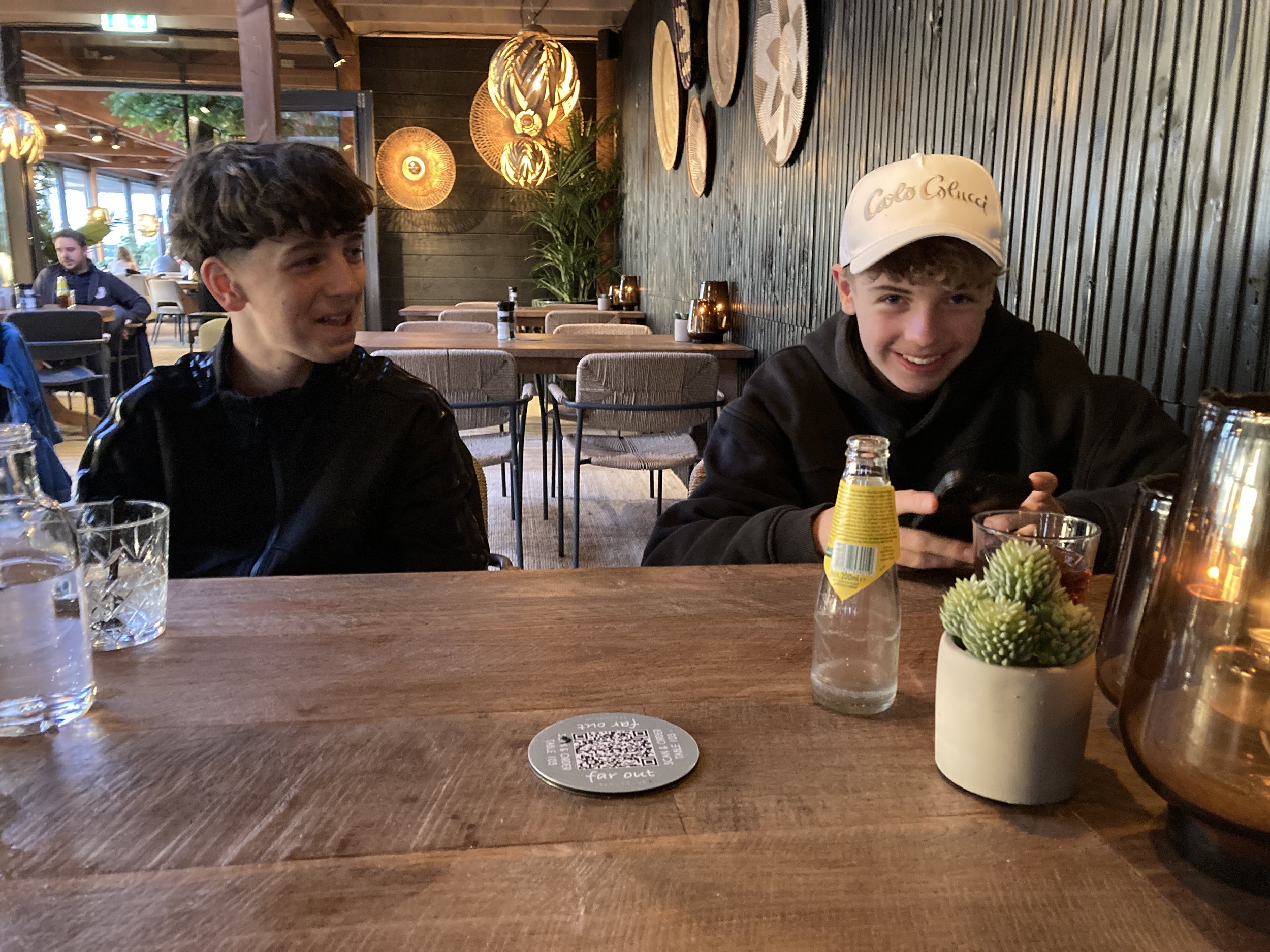 Zwei junge Männer sitzen an einem Holztisch in einem Restaurant, lachen und unterhalten sich. Es gibt Getränke, eine kleine Pflanze und Dekoration auf dem Tisch.