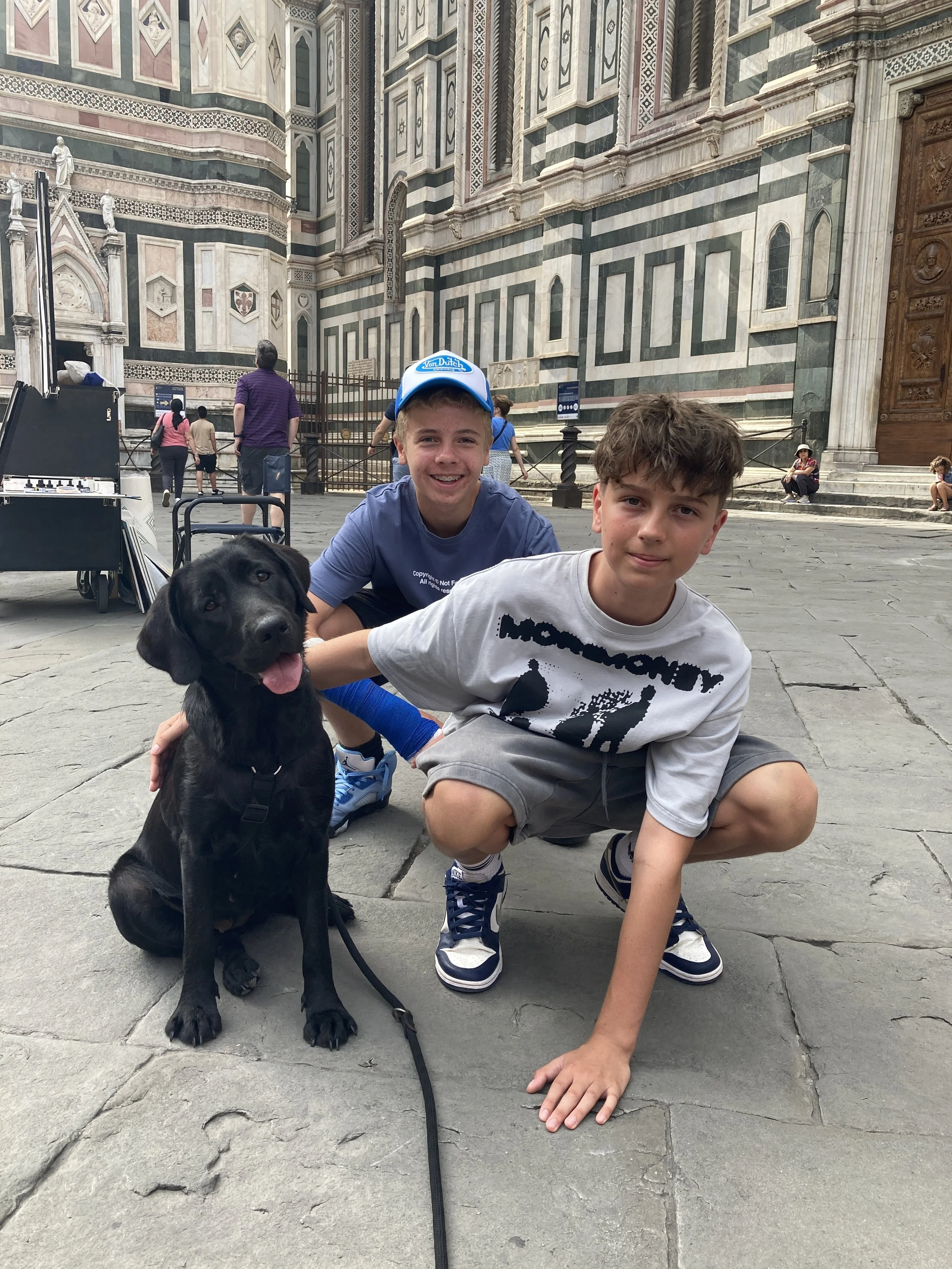 Zwei Jungen und ein schwarzer Hund vor einer historischen Kathedrale, vermutlich in Italien.