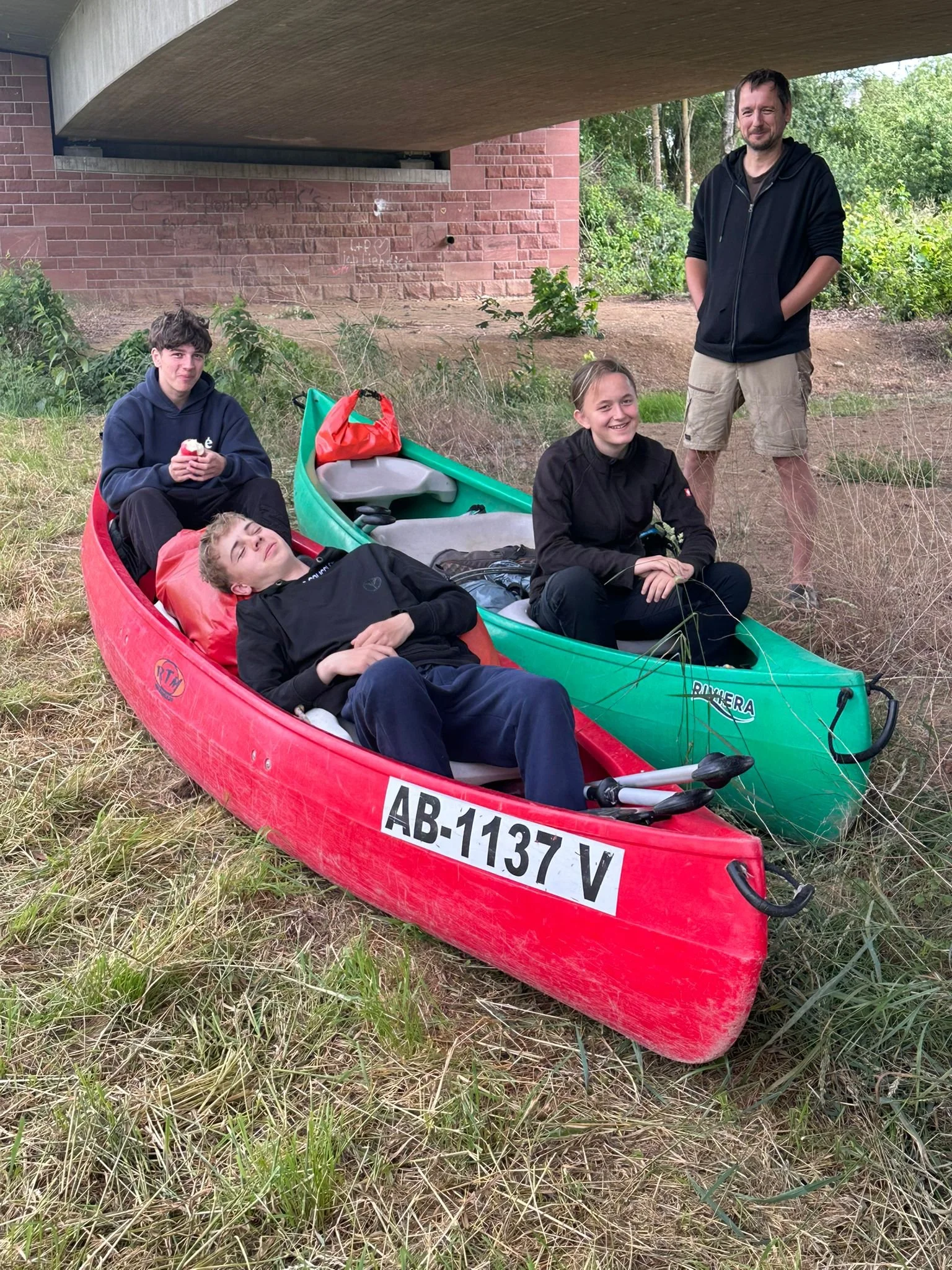 Vier Personen, drei im Wasser und einer stehend, neben zwei Kanus auf dem Gras unter einer Brücke.