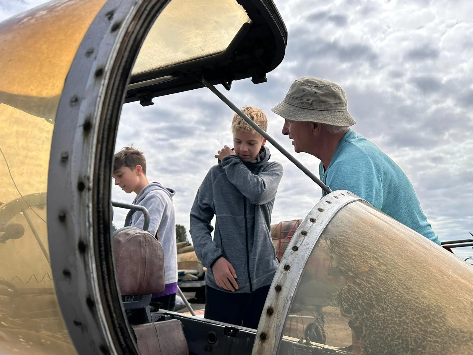 Ein älterer Mann spricht mit zwei jungen Jungen, die neben einem Flugzeug stehen, auf einem Flugfeld unter einem bewölkten Himmel.