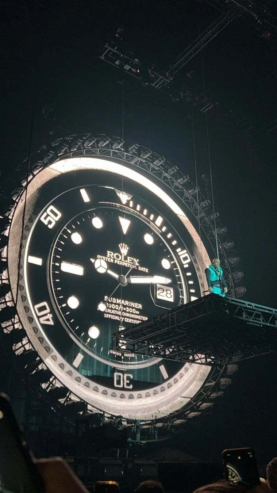 Finn war beim Luciano Konzert in Köln 2024. Großes, leuchtendes Rolex-Uhr-Design im Hintergrund, mit einer Person in Kletterausrüstung auf einer Plattform davor.
