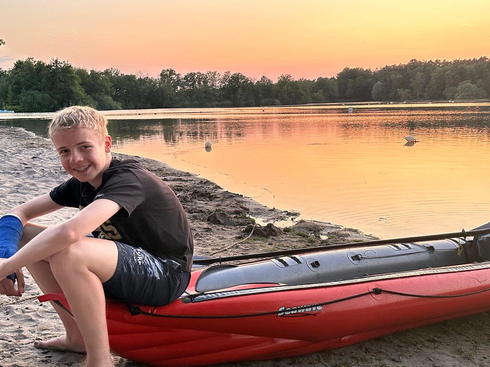 Ein Junge sitzt neben einem roten Kajak am Seeufer bei Sonnenuntergang, umgeben von Wasser und Bäumen.