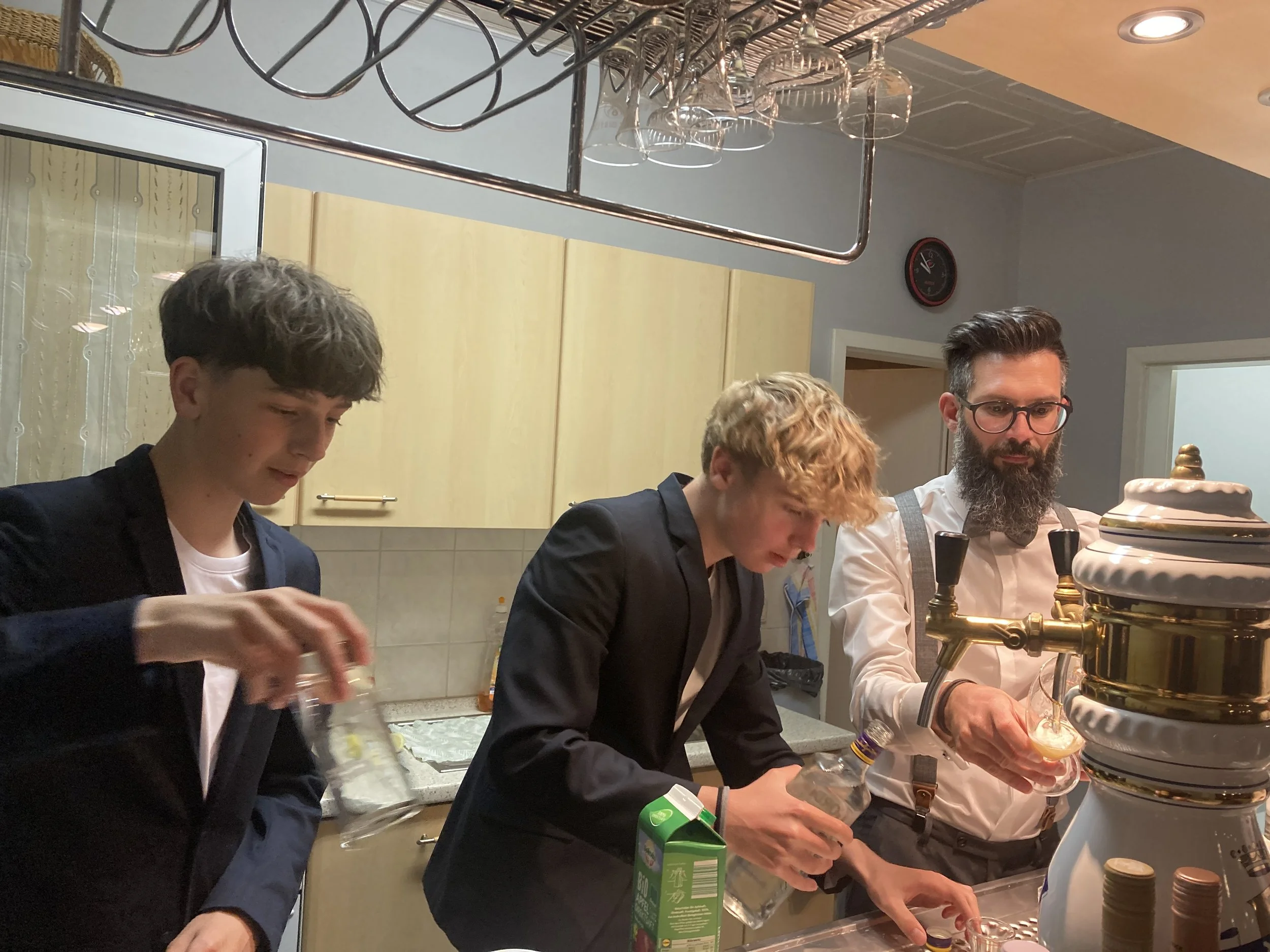 Drei Männer in formeller Kleidung bereiten Getränke an einer Bar vor, während eine Person einen Getränkespender bedient.