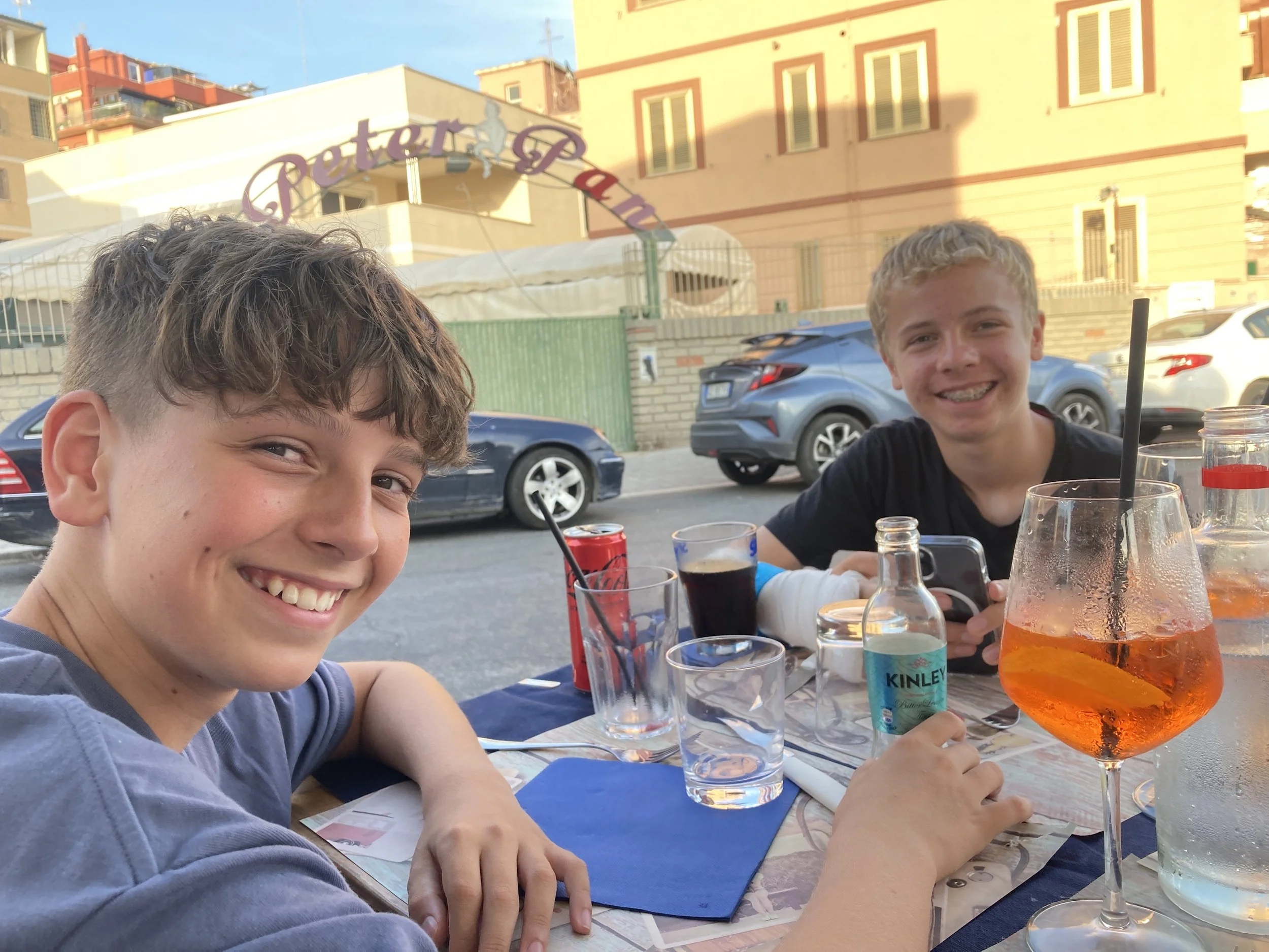 Zwei Jungen sitzen an einem Restauranttisch im Freien, lächeln in die Kamera, mit Getränken und einem Smartphone, im Hintergrund Autos und ein Gebäude bei Sonnenuntergang.