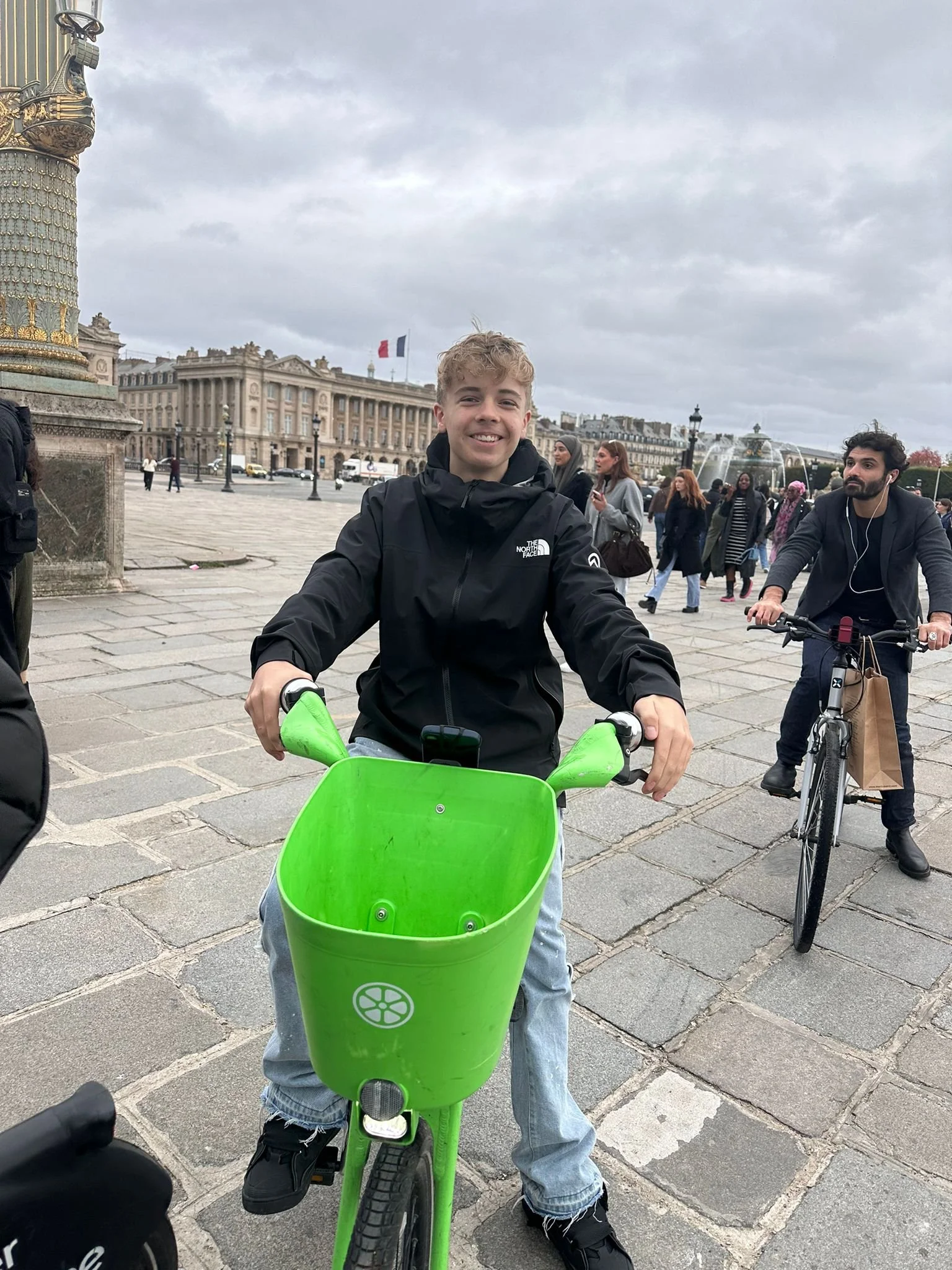 Ein Junge fährt auf einem grünen Fahrrad in einer Stadt, im Hintergrund sind mehrere Menschen und ein Gebäude mit französischer Flagge, es ist cloudy.