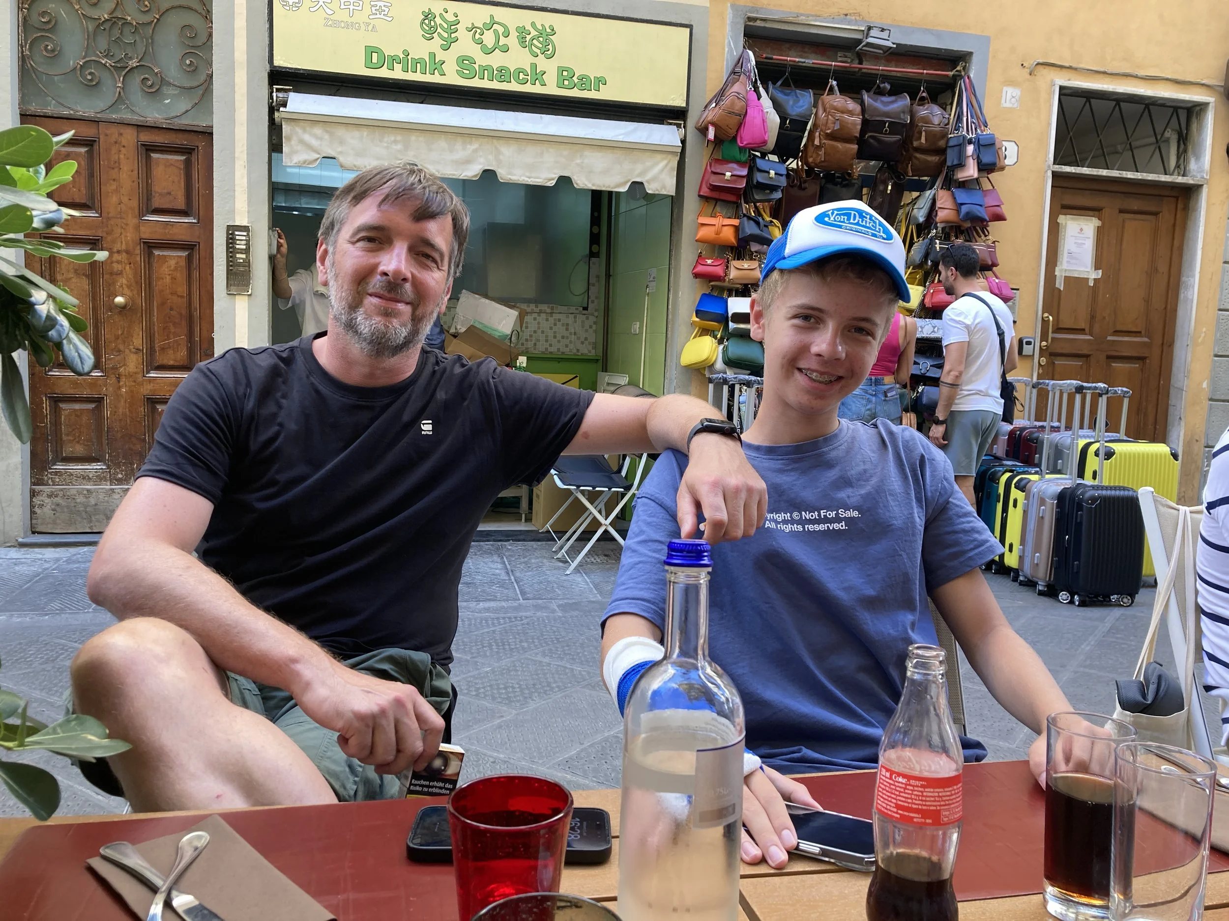 Zwei Männer, einer älter mit grauem Bart und einer jüngere mit Brille, sitzen an einem Tisch in einem Cafè oder Restaurant im Freien. Im Hintergrund ist eine Tasche- und Koffer-Verkaufstheke. Sie sind lächelnd und haben Getränke vor sich auf dem Tisc