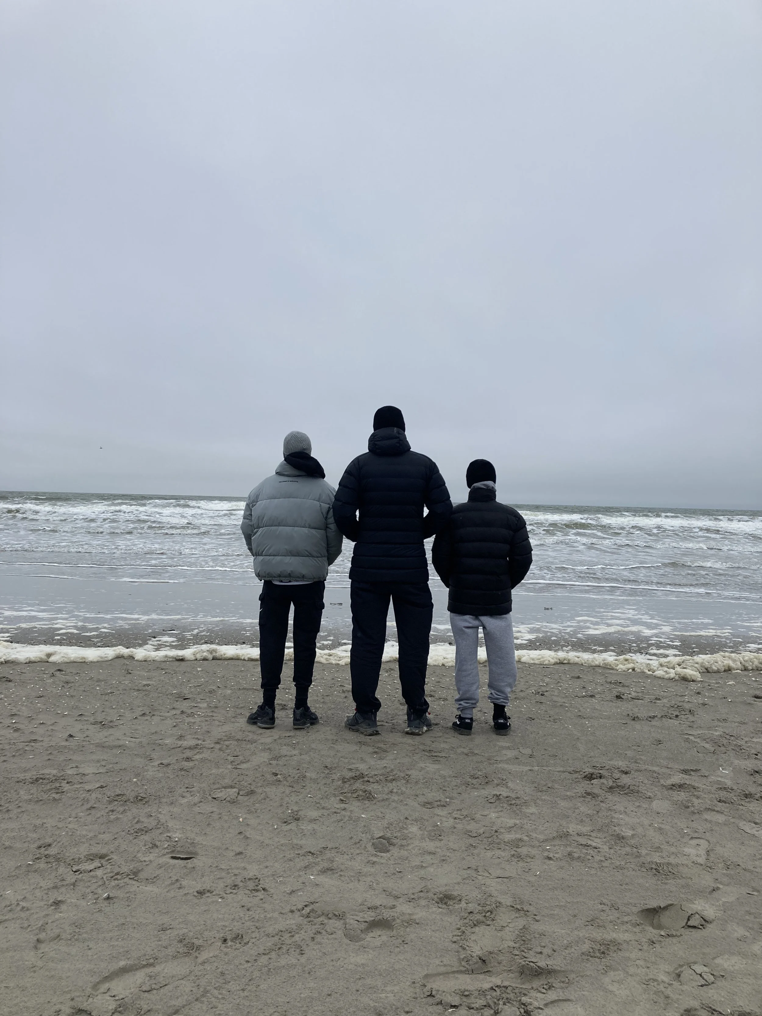 Drei Personen stehen am Strand und schauen aufs Meer, während bewölkter Himmel die Atmosphäre prägt.