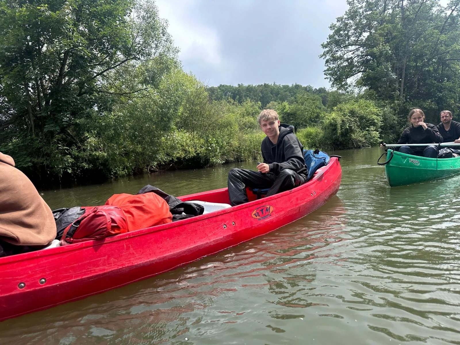 Drei Personen paddeln in Kanus auf einem Fluss, umgeben von Bäumen. Einer lächelt in die Kamera, die anderen sitzen und genießen die Natur.