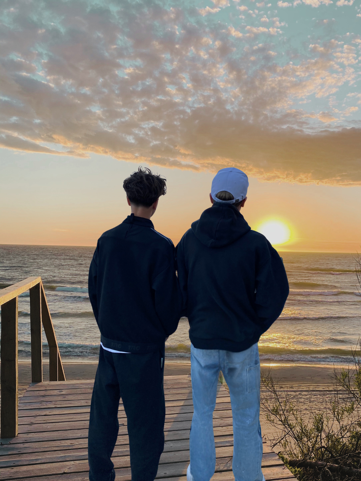 Zwei Brüder (Finn und Jan) schauen bei Sonnenuntergang aufs Meer, stehen auf einem Holzsteg am Strand in Holland Zandvoort, tragen dunkle Kapuzenjacken und lässige Hosen, im Hintergrund sind Wolken und der Himmel in warmen Farben