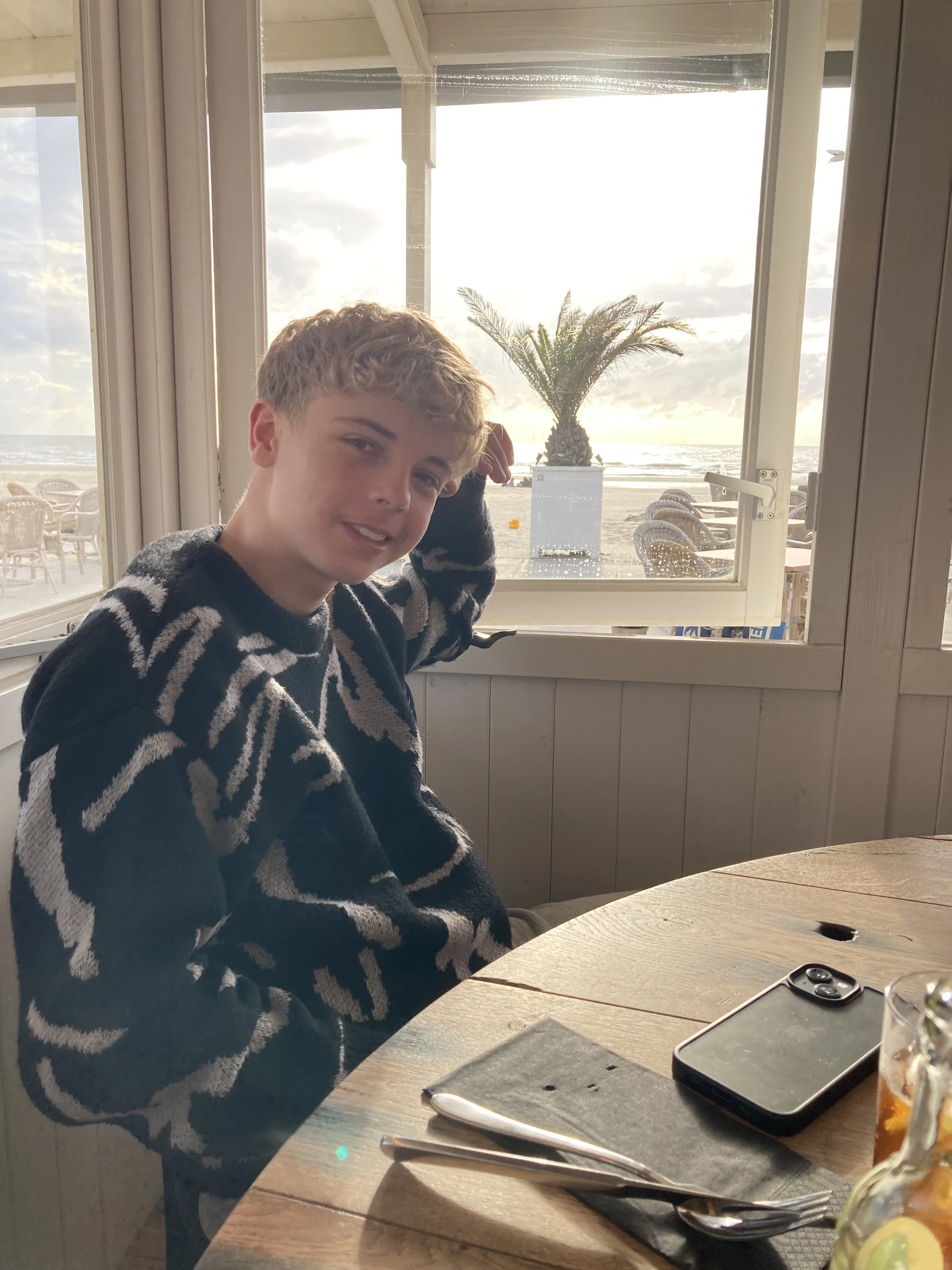 Ein Junge sitzt an einem runden Holztisch in einem Restaurant, in der Nähe eines Fensters mit Blick auf den Strand, der bei Sonnenuntergang zeigt, mit einem Palmen im Hintergrund.