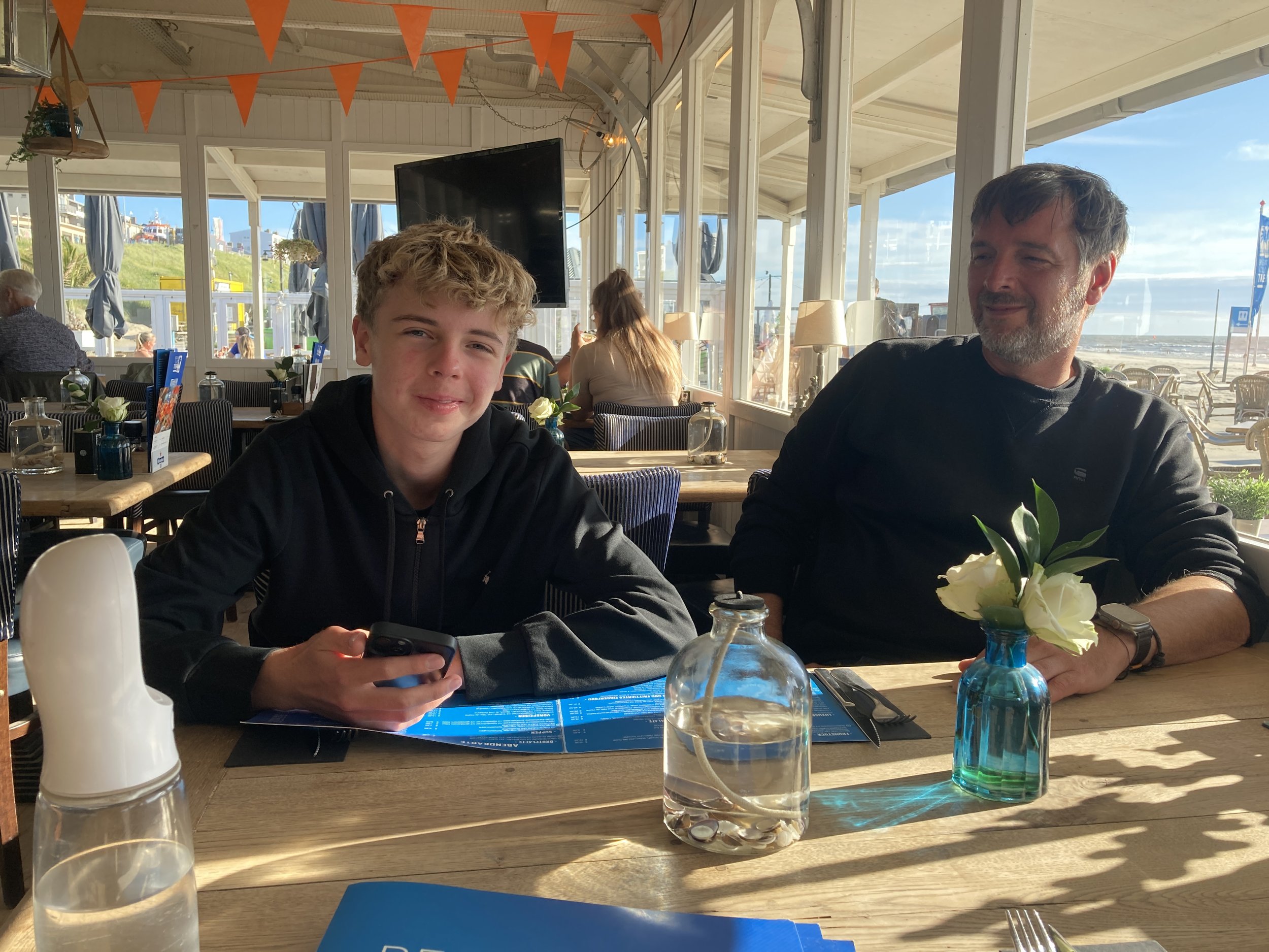 Zwei Männer sitzen zusammen in einem Strandrestaurant. Der jüngere Mann hält ein Smartphone in der Hand, während der ältere Mann eine kleine Vase mit weißen Blumen hält. Im Hintergrund sind Fenster mit Blick auf den Strand und das Meer, sowie andere 