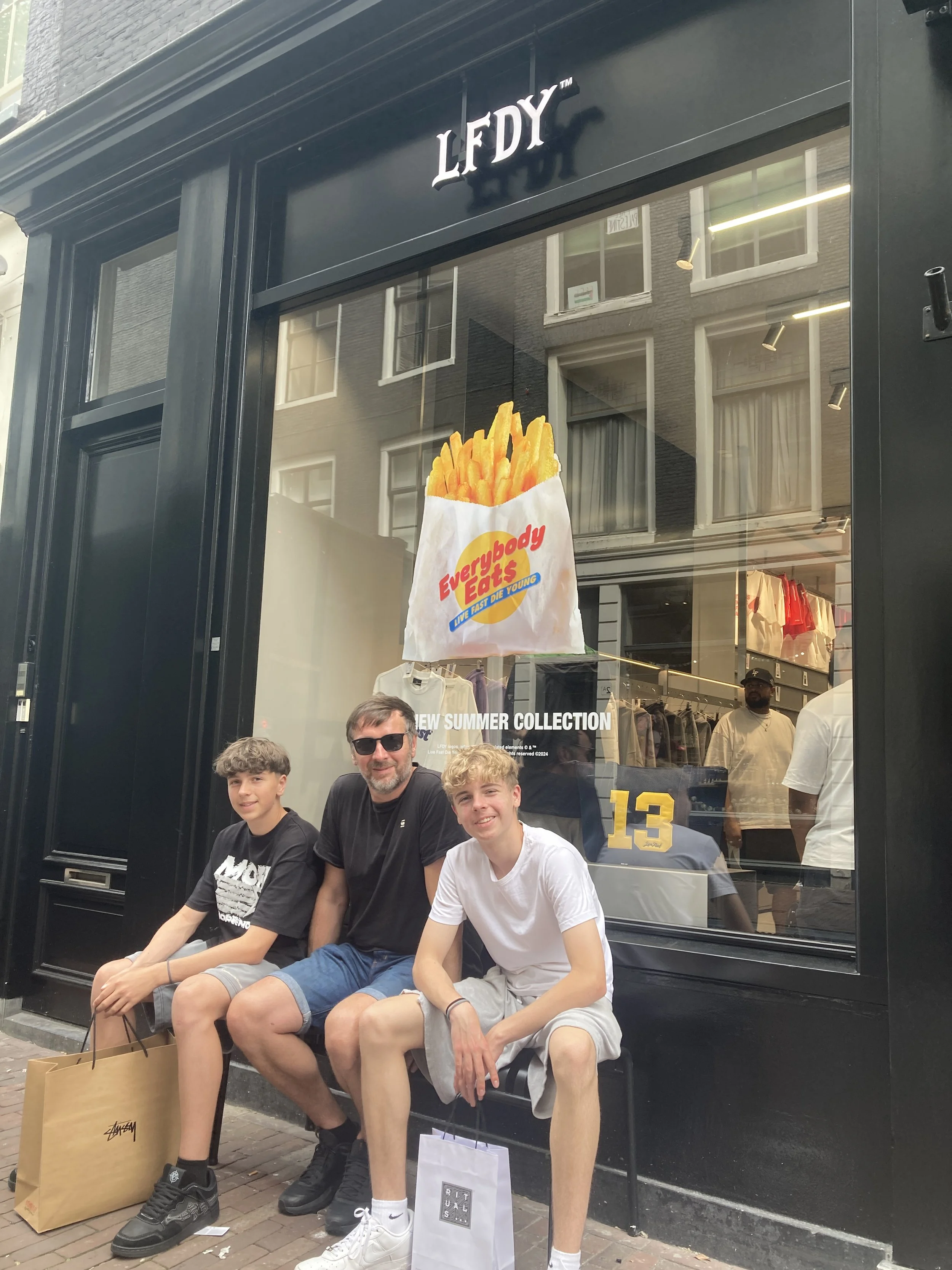 Drei Jungs und ein Mann sitzen vor einem Geschäft mit großen Fenstern, das eine modische Kleidungskollektion bewirbt. Hinter ihnen hängt ein großes Plakat mit einem Pommes-Frites-Beutel, der die Marke 'Everybody Eats' zeigt.