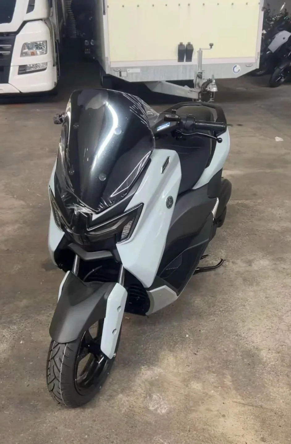 Finns Roller. Sein Traum. Den hätte er zu seinem 16. Geburtstag bekommen. Ein weißes Elektromotorrad mit schwarzem Windschutz und schwarzem Sitz, parkend auf Betonboden, im Hintergrund ein Lastwagen und andere Motorräder.