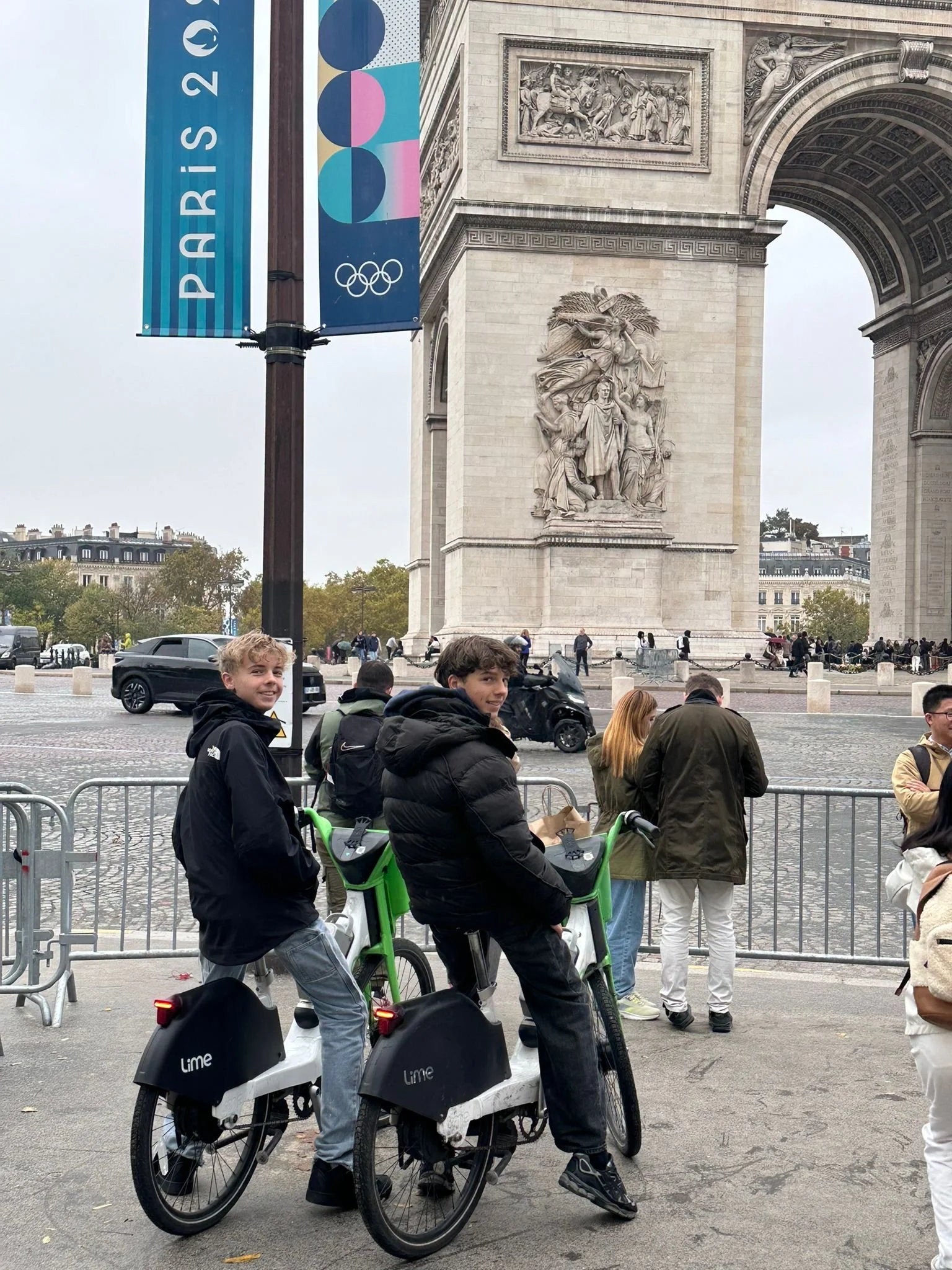 Zwei junge Männer stehen mit E-Bikes in Paris vor dem Arc de Triomphe, umgeben von Touristen, bei bedecktem Himmel.