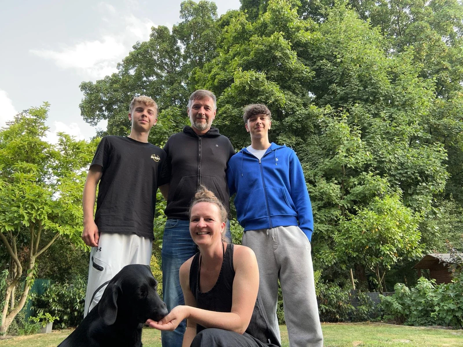 Eine Familie aus fünf Personen, inklusive eines Hundes, steht in einem Garten mit grünen Bäumen im Hintergrund, lächelt und posiert für ein Foto.