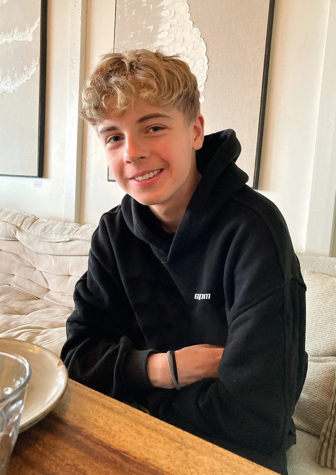 Ein lächelnder junger Mann Finn mit blonden, lockigen Haaren trägt einen schwarzen 6pm-Hoodie und sitzt an einem Tisch in einem gemütlichen Raum mit beigefarbenen Sitzmöbeln und moderner Wanddekoration.