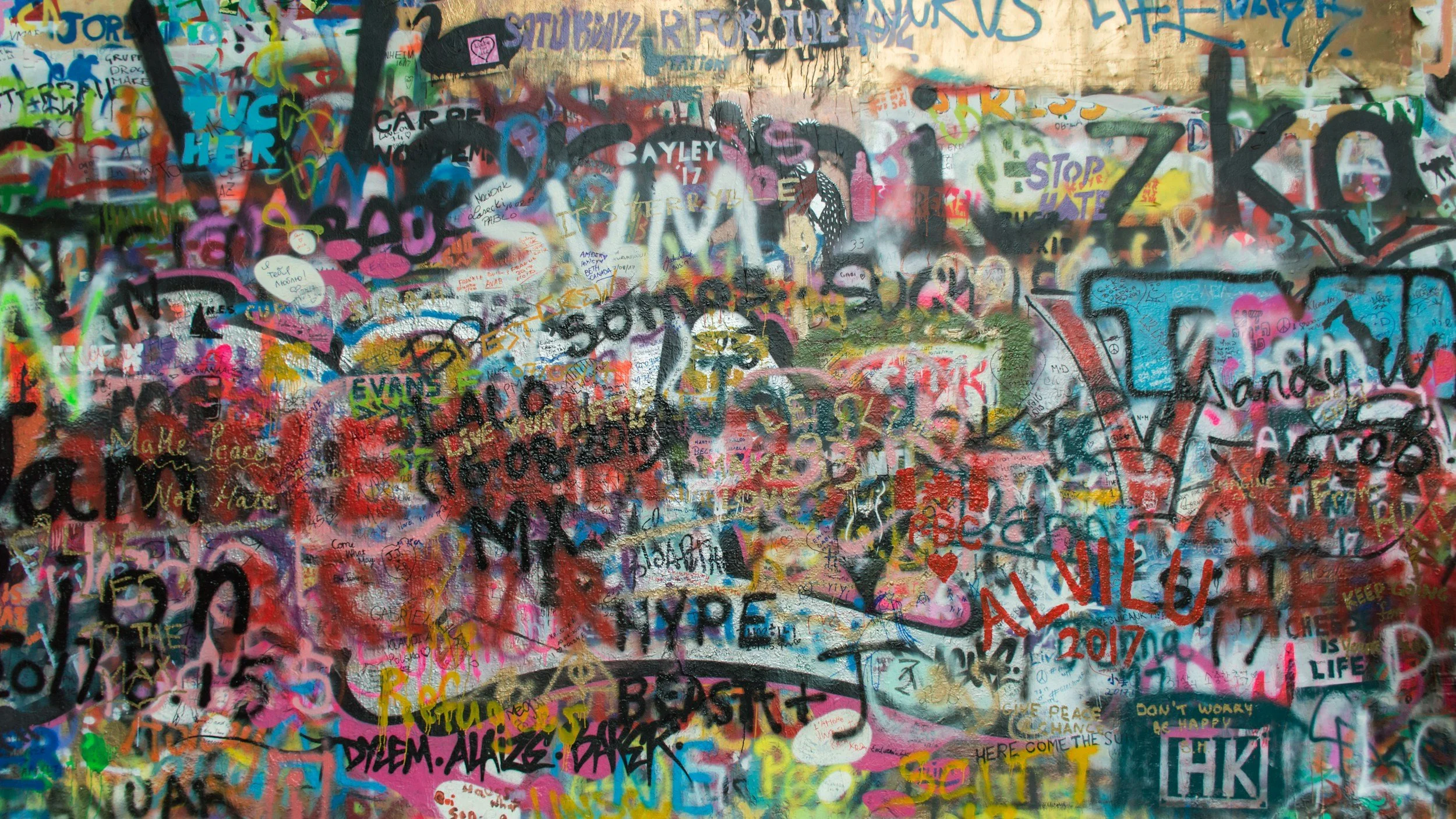 A colorful wall covered in overlapping graffiti, spray paint, and various writings including words, phrases, and signatures in multiple colors and styles.