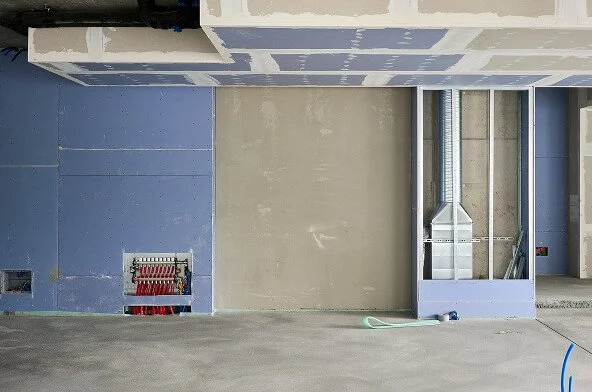 Construction site with blue drywall, a gray concrete wall, and an unfinished duct system with metal ducts and a flexible duct connected to a white box.